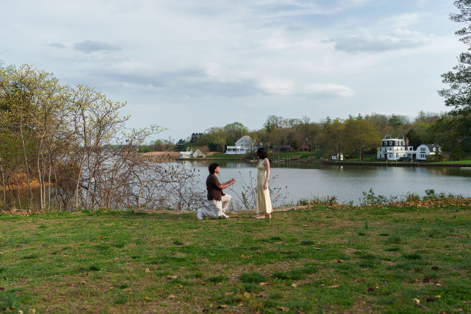 Man proposing to woman on grassy riverbank with calm water and trees in background.