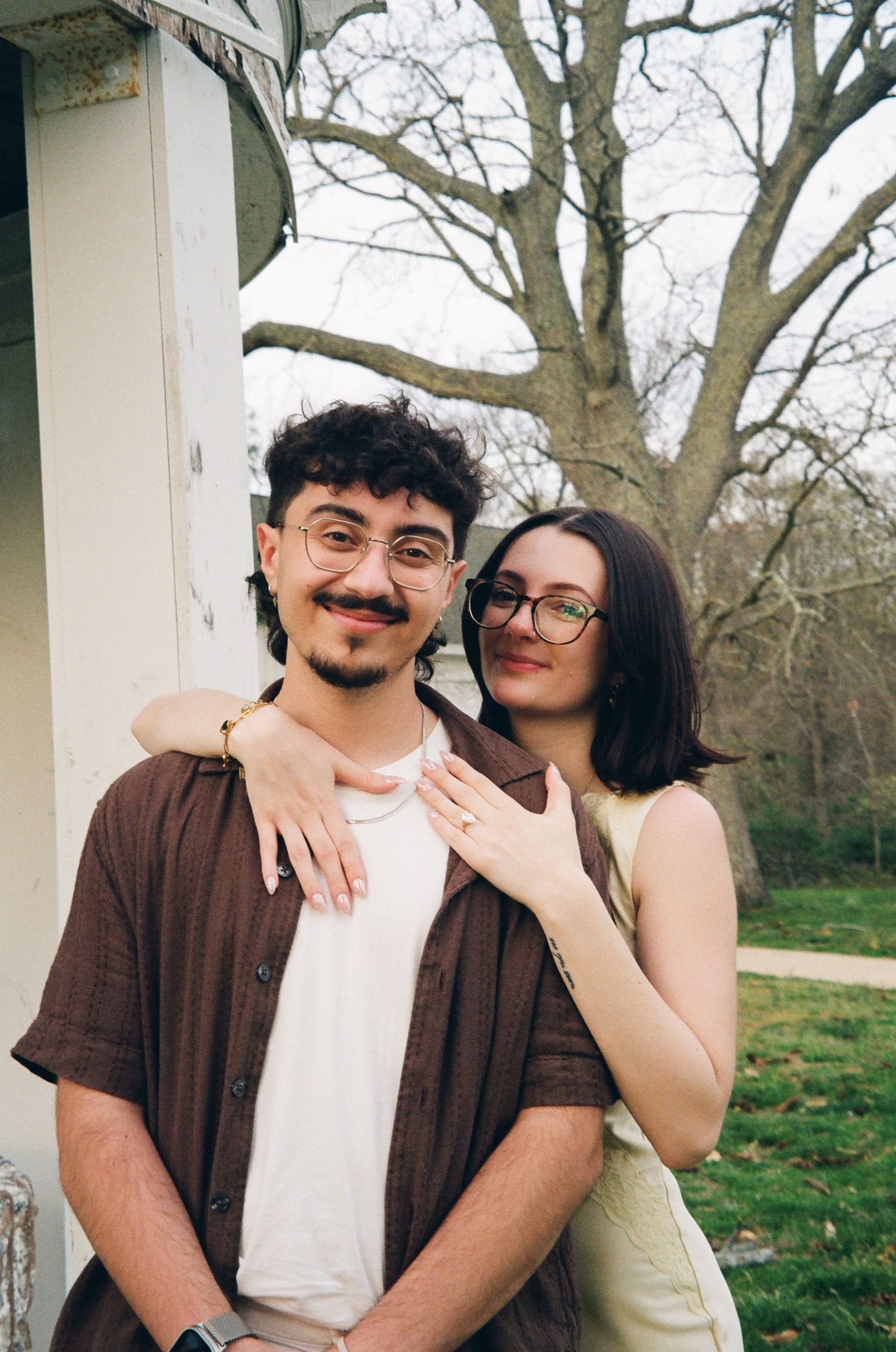 Couple smiling near white columns outdoors, woman in yellow dress embracing man in brown shirt.