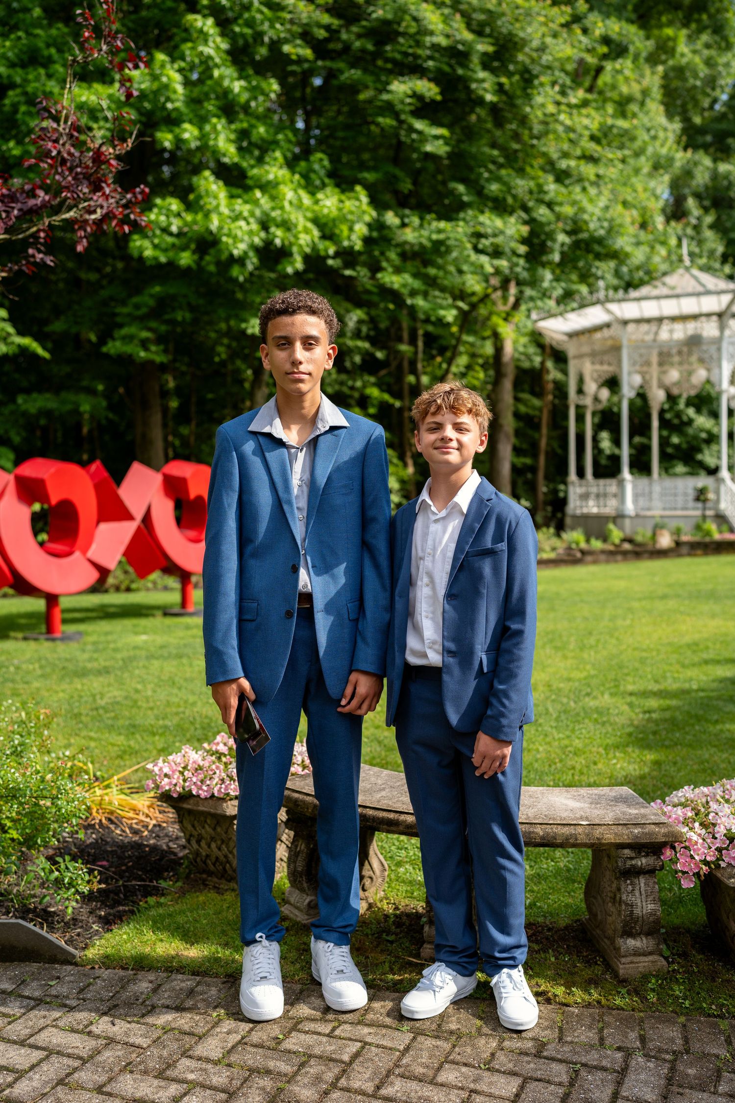 Two boys in matching blue suits posing in a garden with a gazebo backdrop.
