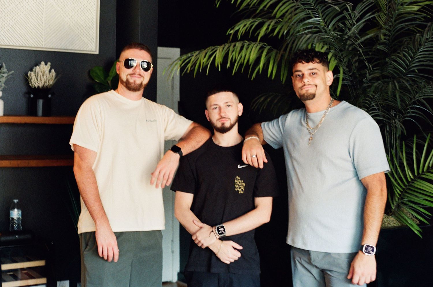 Three young men posing together indoors near tropical plants in a modern dark-themed interior space.