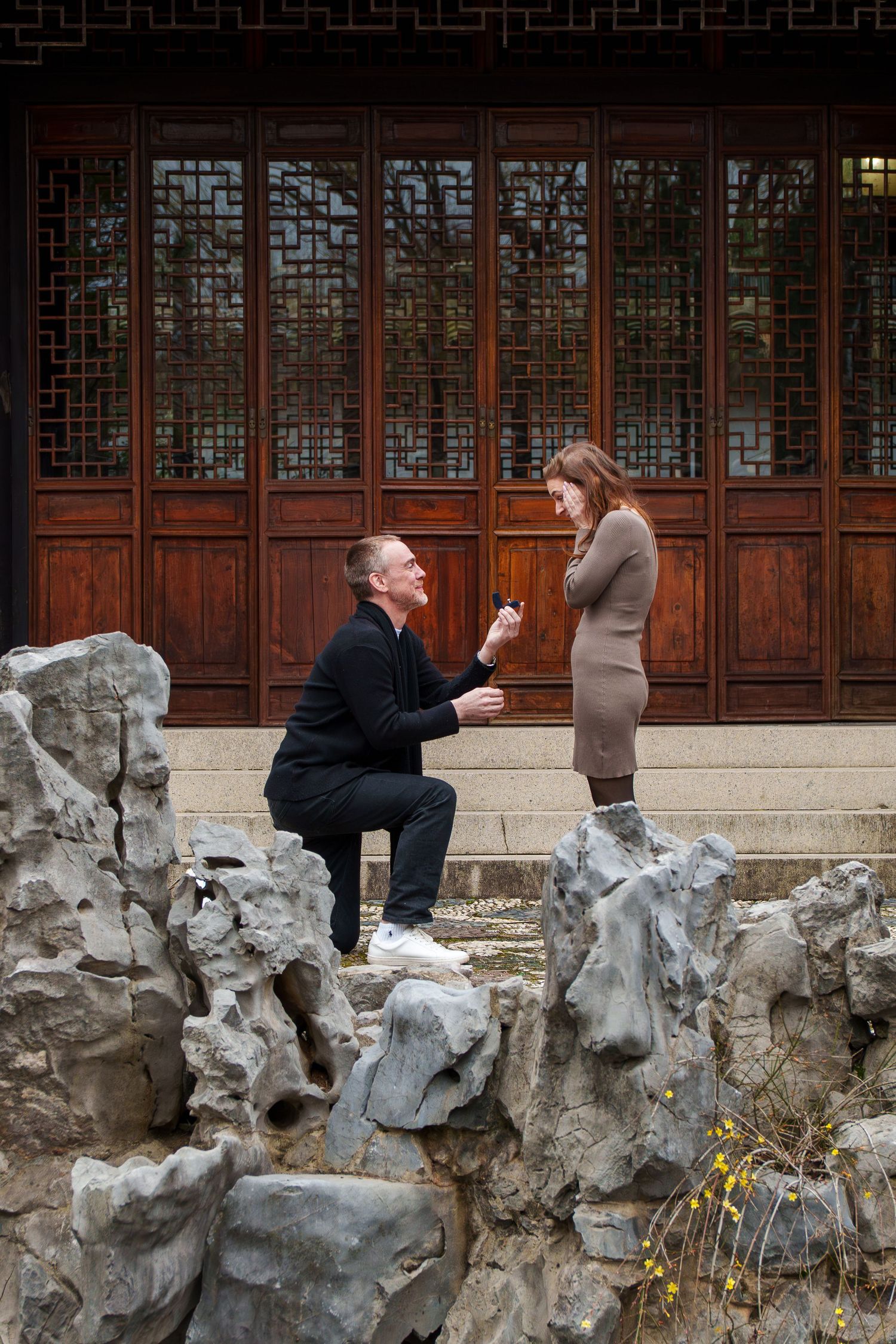 Man proposing on one knee to woman on rocky landscape before a Chinese pavilion.