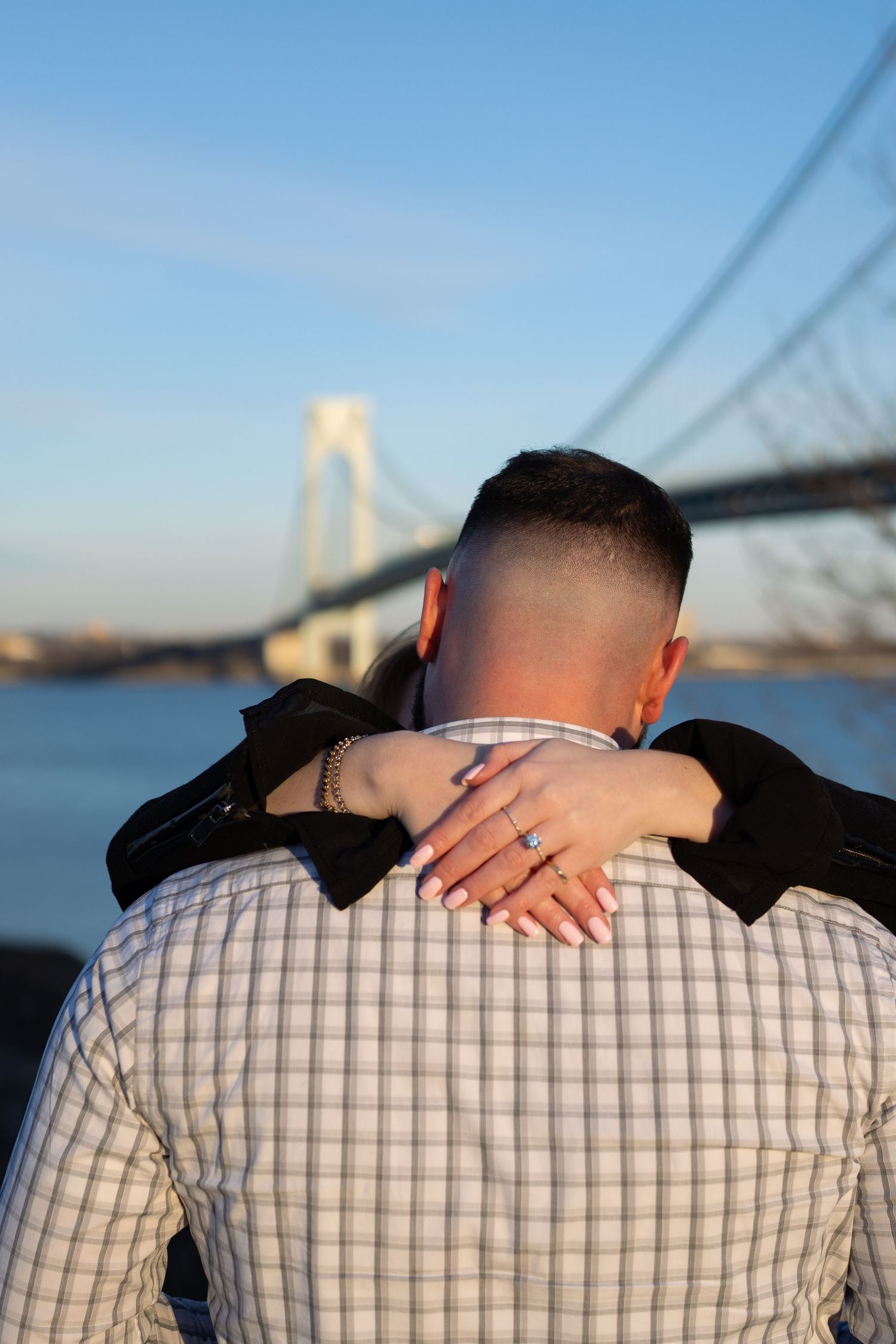 Woman wrapping arms around man's neck showing off diamond engagement ring with bridge backdrop.
