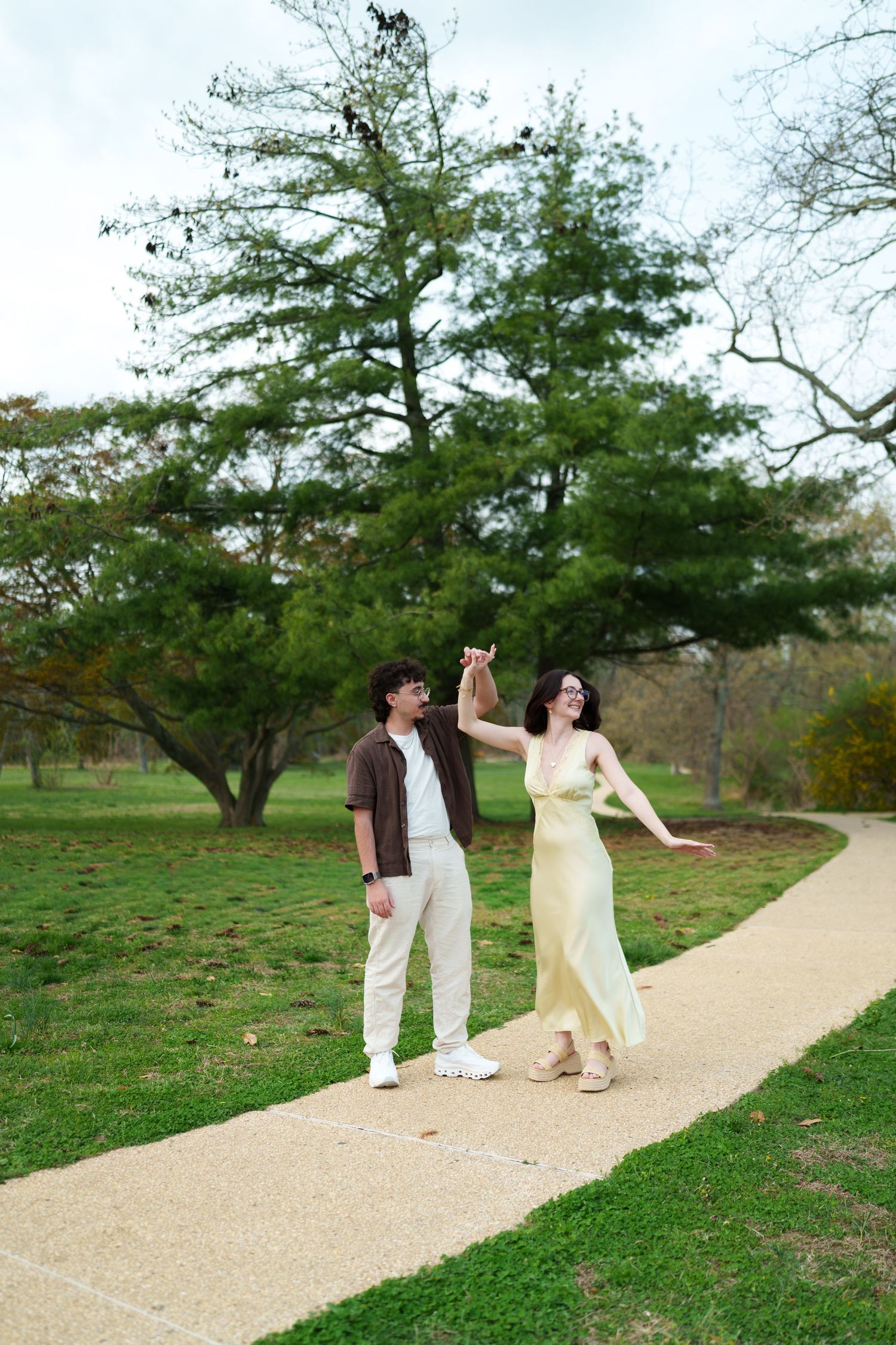 Happy couple dancing and holding hands on a scenic park pathway with green trees.