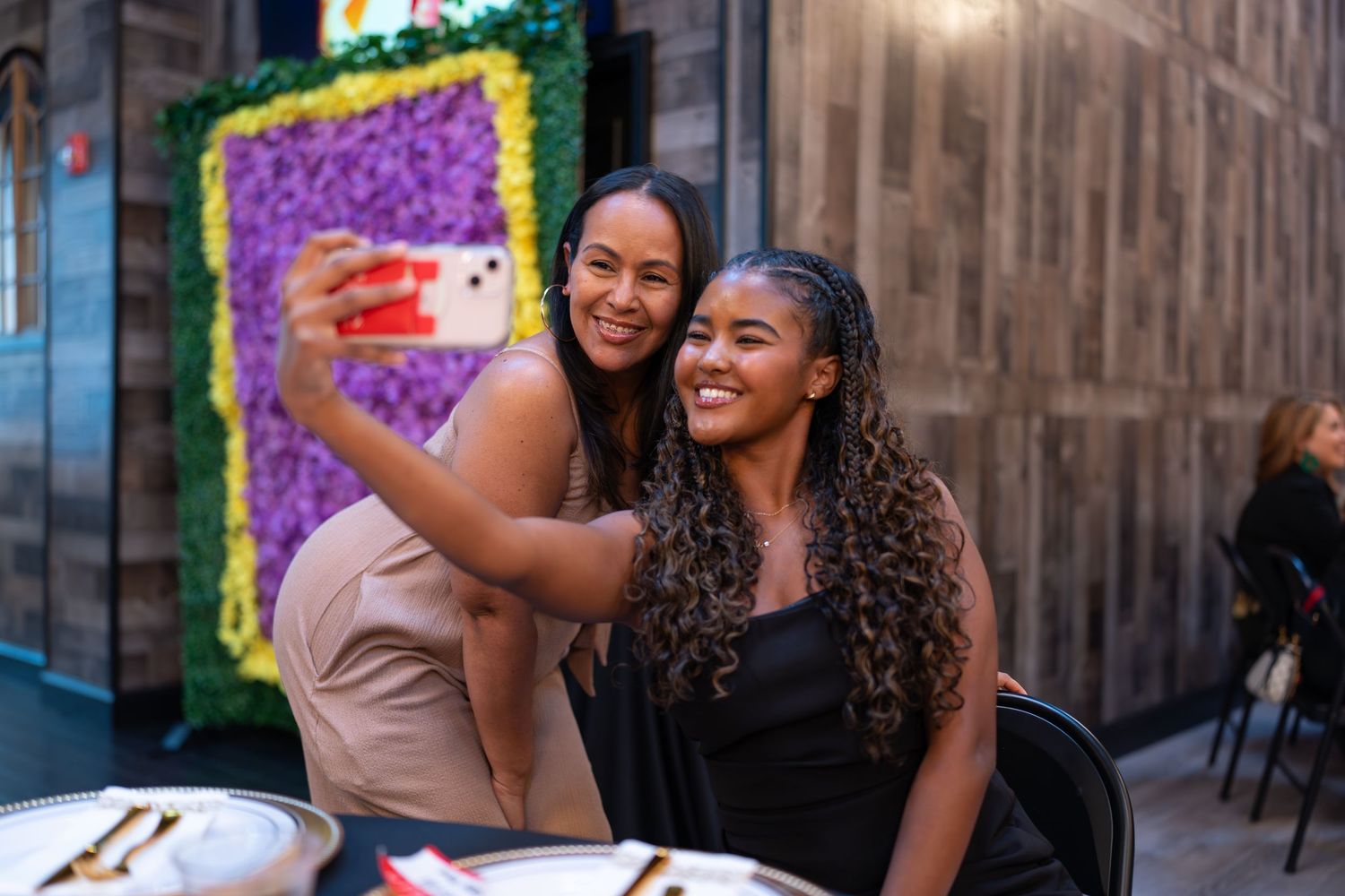 Two young women taking a selfie together at a table with a floral wall backdrop.