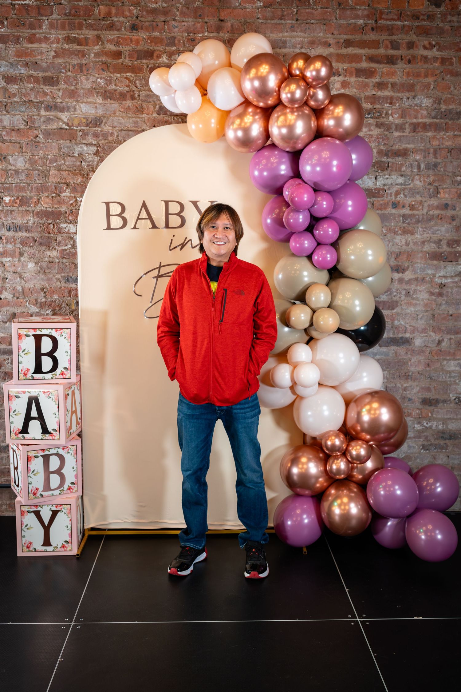Man in red jacket smiles at baby shower with balloon arch and BABY letter blocks backdrop.