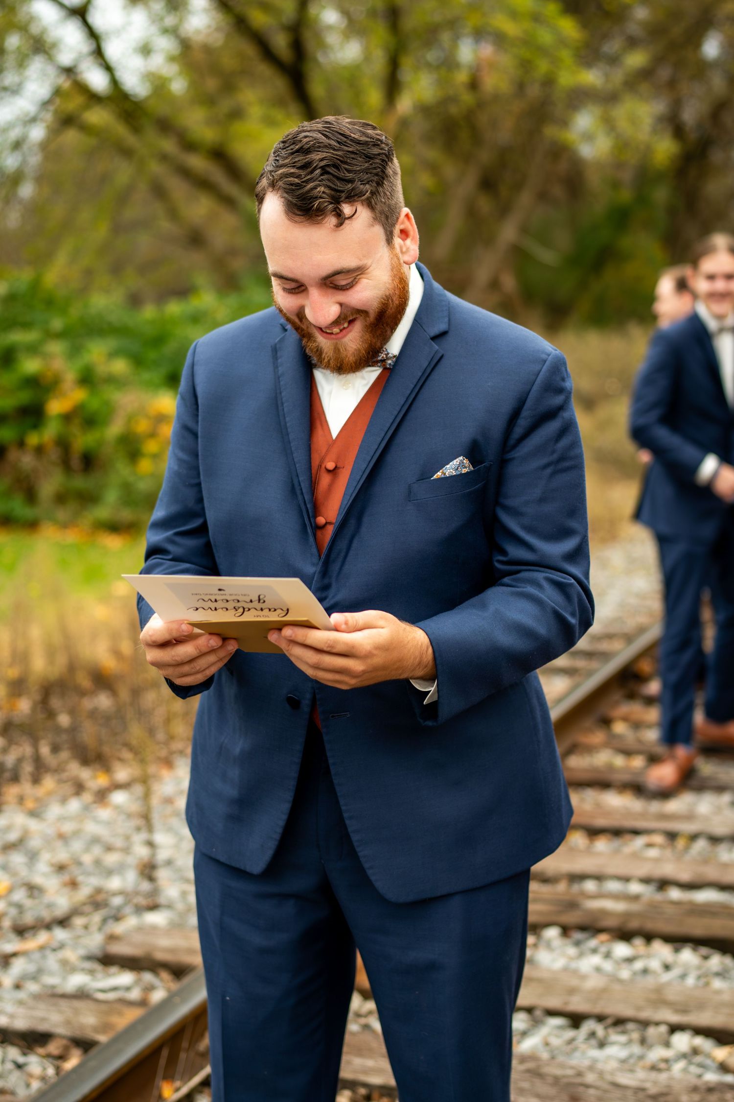 Smiling groom in navy suit reads a card on railroad tracks at his autumn wedding.