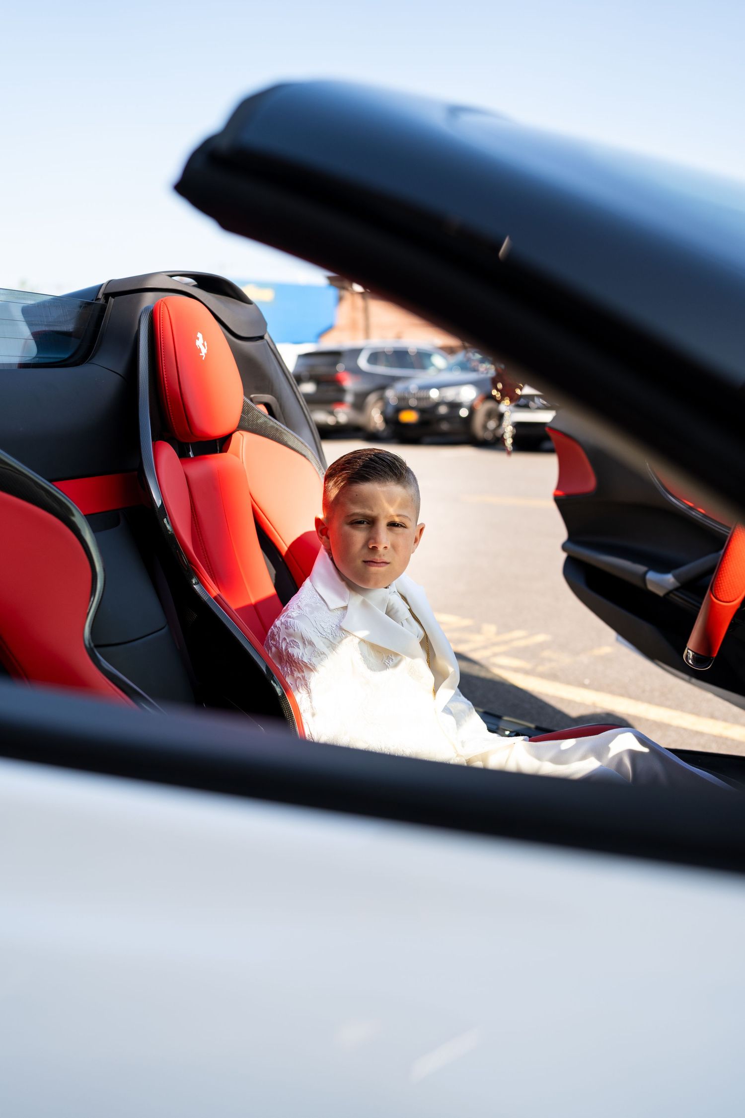Young boy in white suit sitting in red interior Ferrari convertible.