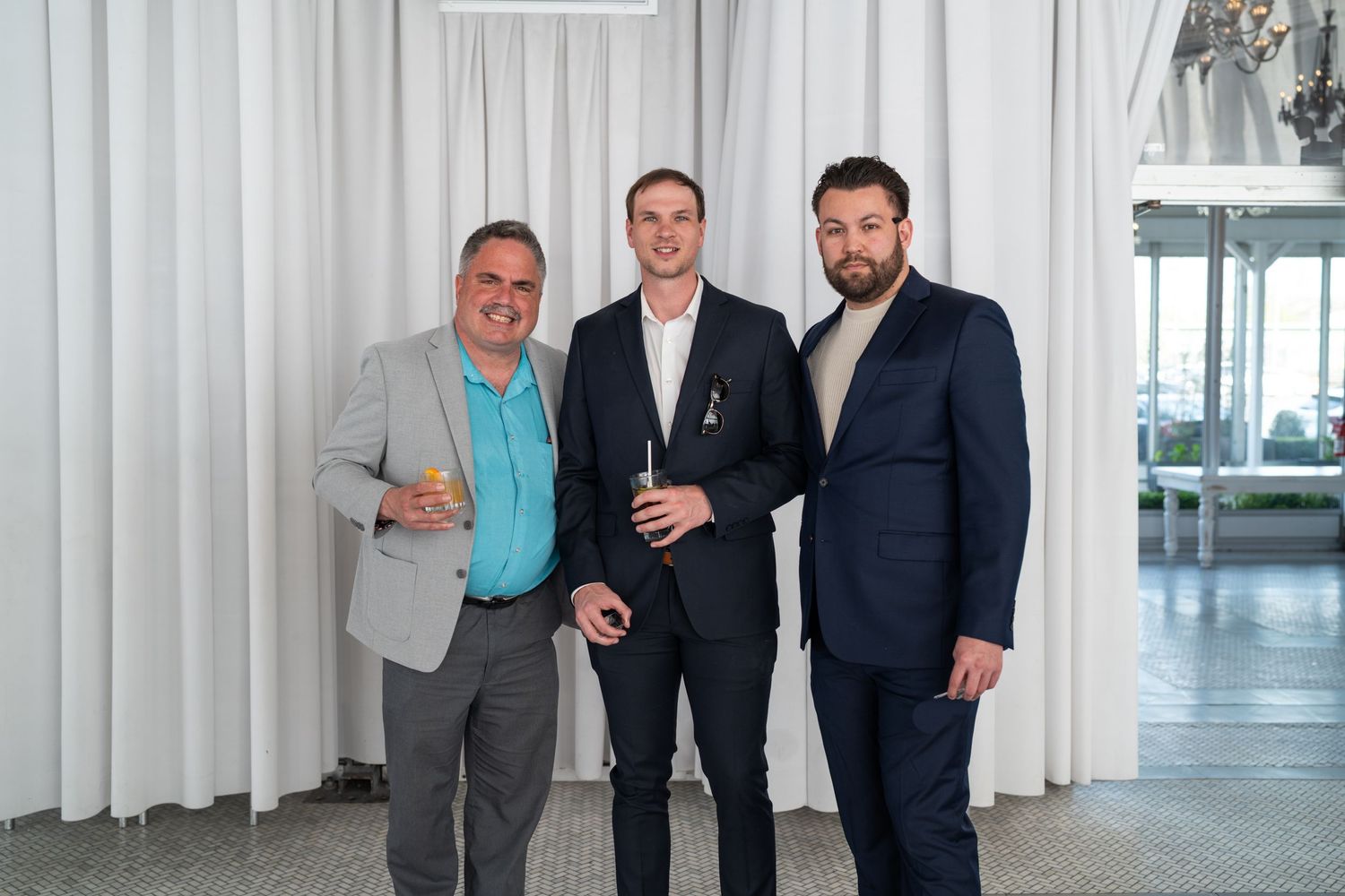 Three men in suits pose together at an elegant white curtained event venue.