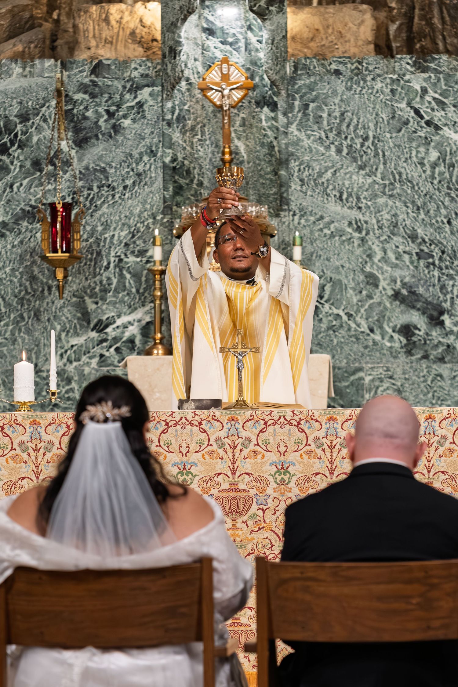 Priest elevating the Eucharist at the altar while bride and groom kneel during Catholic wedding Mass.