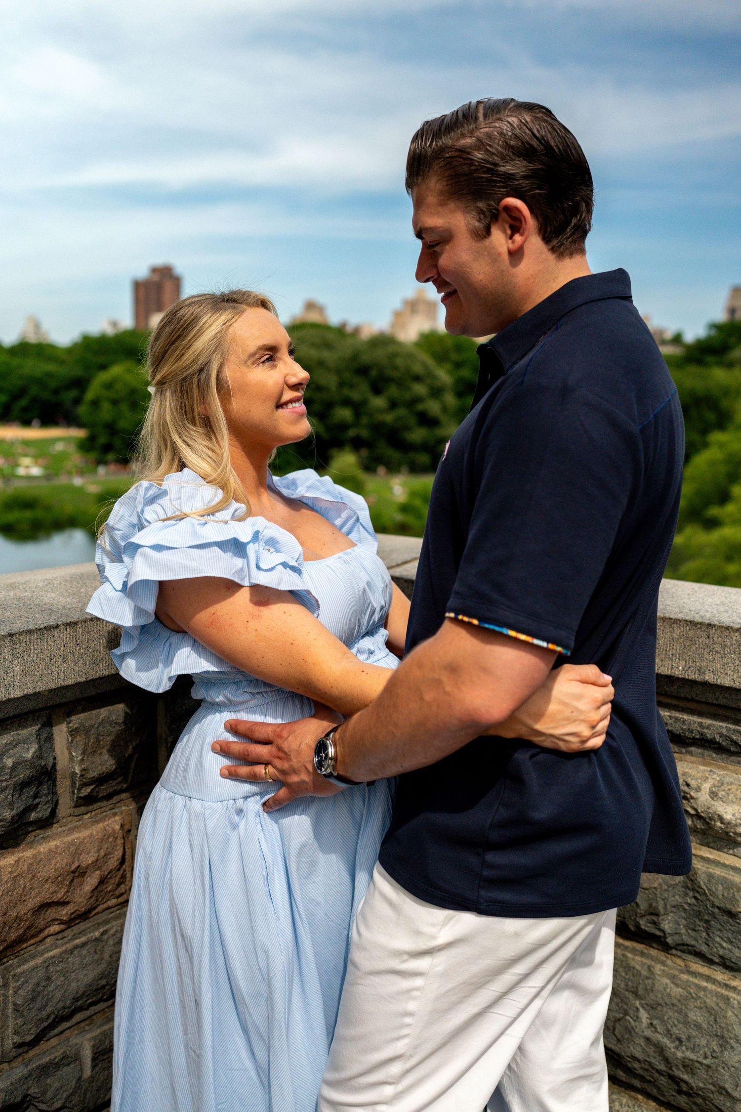 Happy couple embracing on a stone bridge in Central Park with city skyline behind.