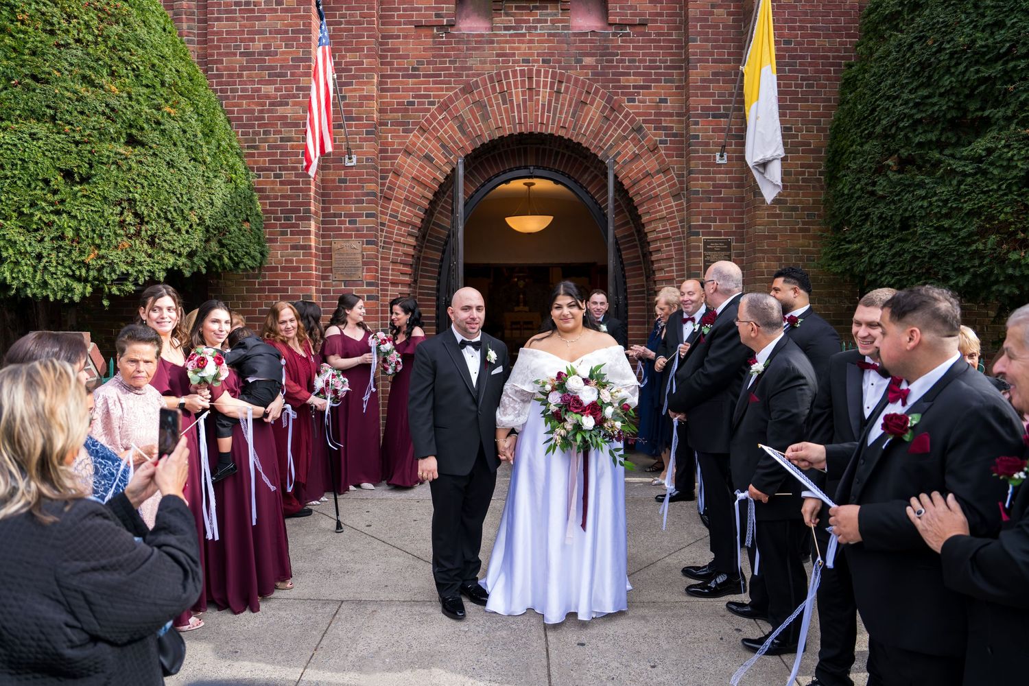 Bride and groom exit brick church as wedding party lines up in burgundy attire for celebration.
