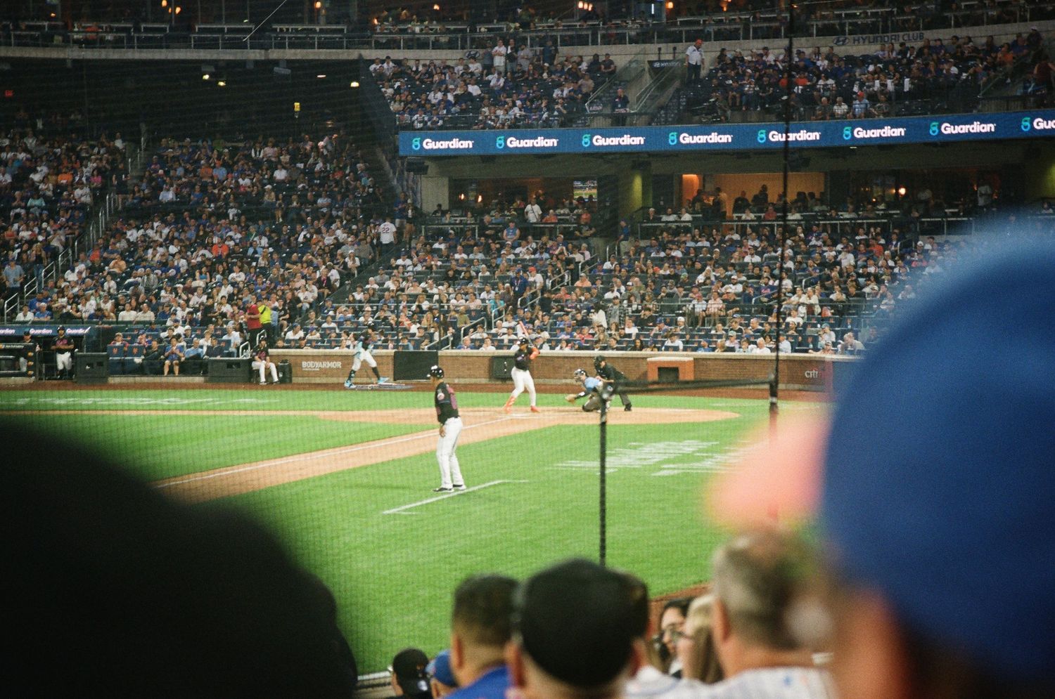 Packed baseball stadium crowd watches a night game from the lower field-level seats.