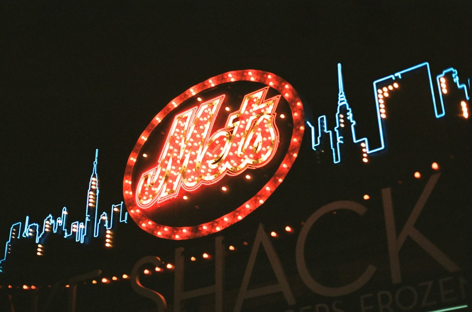 Illuminated New York Mets neon sign with NYC skyline at night outside Citi Field.