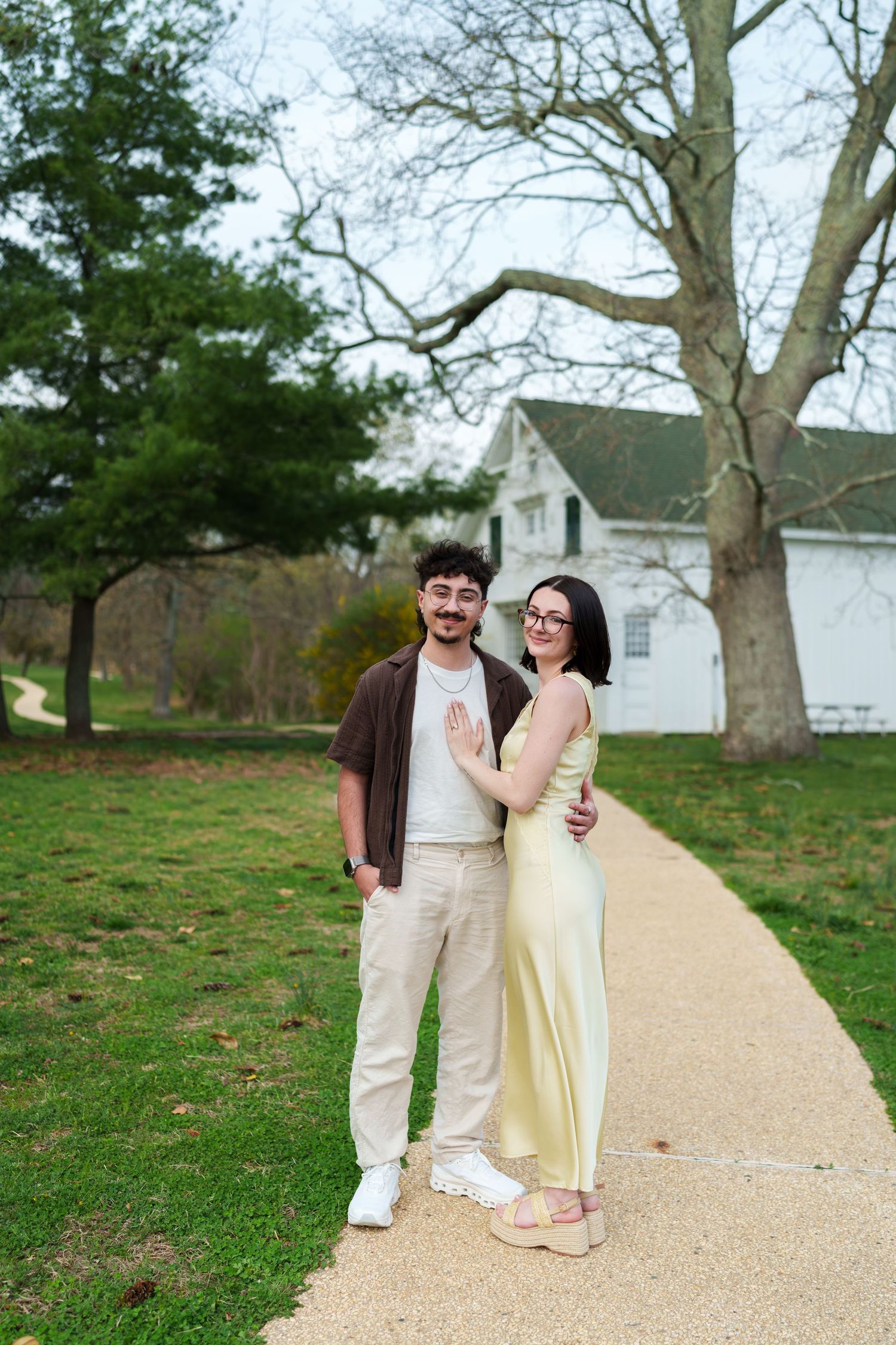 Smiling engaged couple posing on a garden path with a white historic building behind them.
