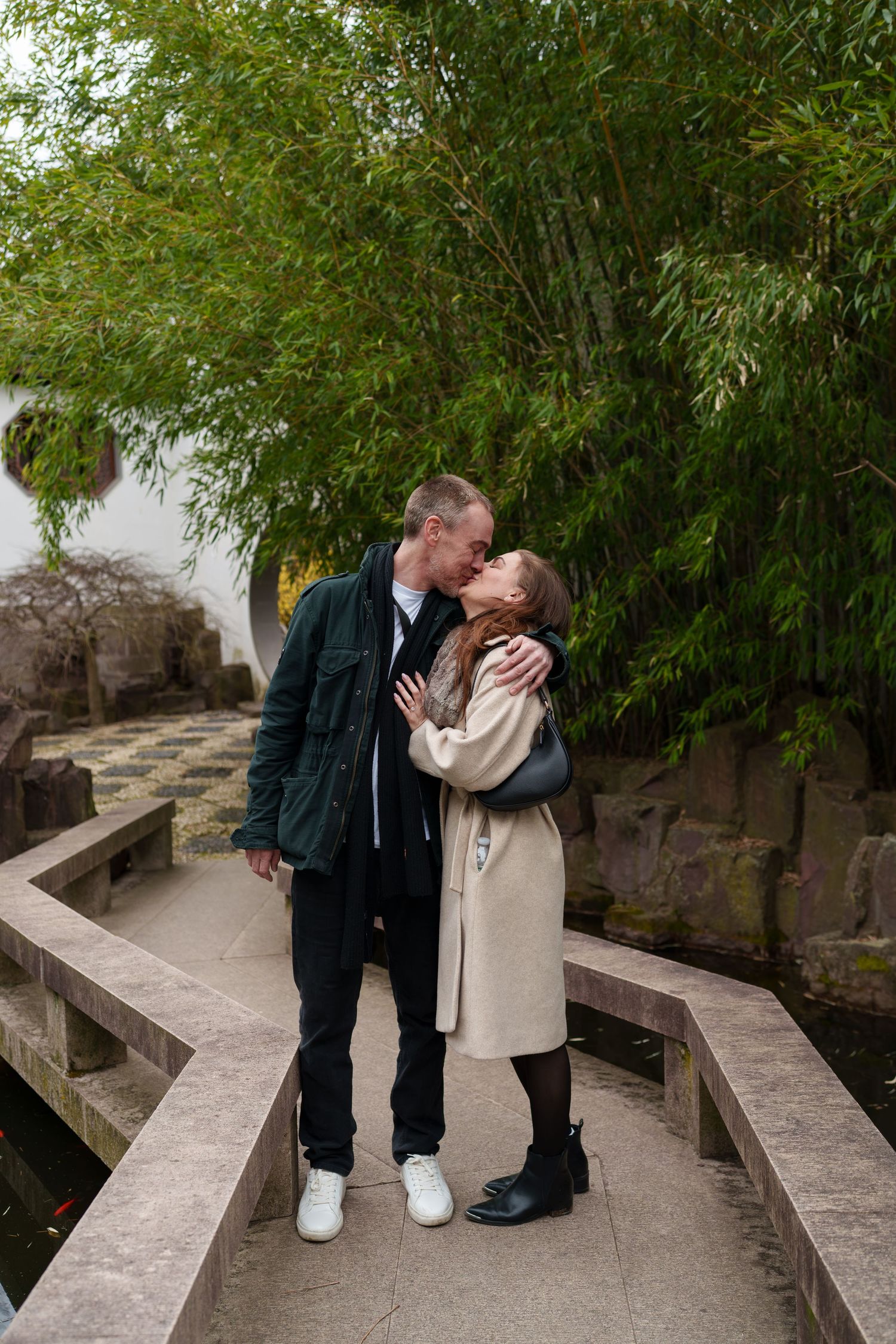 Couple sharing a romantic kiss on a stone bridge surrounded by lush bamboo in a serene garden.