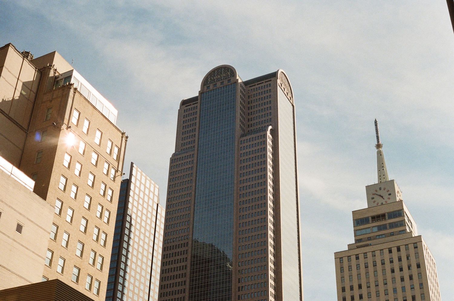 Downtown Dallas skyline featuring modern glass skyscrapers and historic buildings against a bright blue sky.