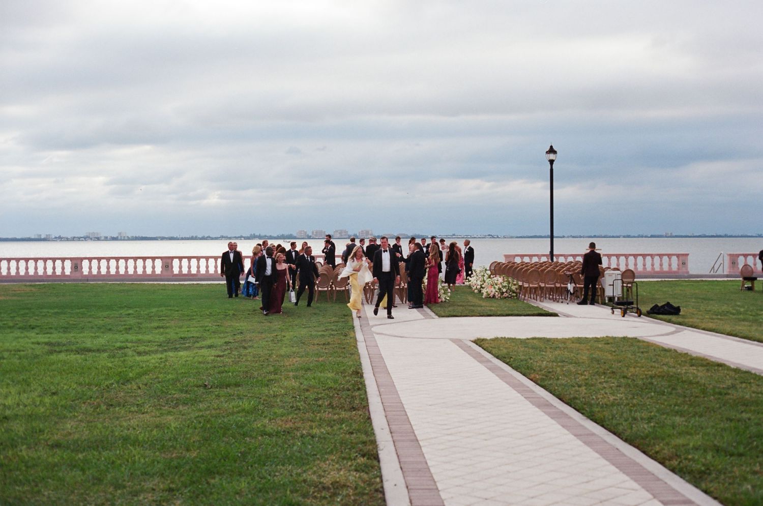 Wedding guests gathered on waterfront lawn with scenic bay views under overcast skies.