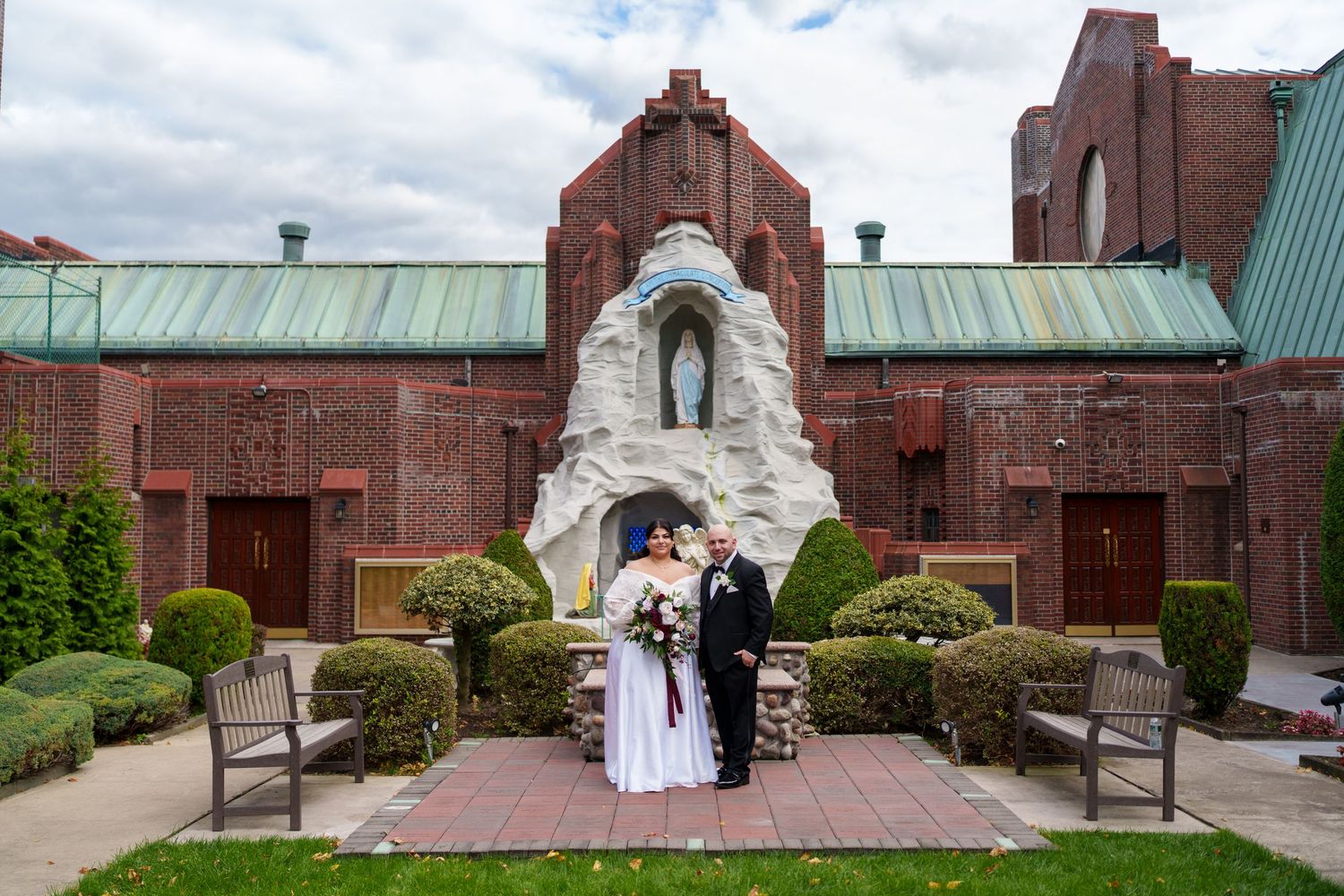 Wedding couple poses before a grand brick church with a white grotto statue and manicured hedges.