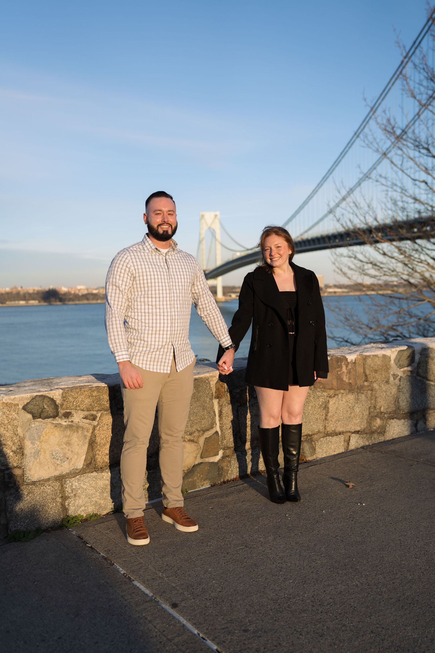 Newly engaged couple holding hands and smiling beside the Verrazzano-Narrows Bridge at sunset.