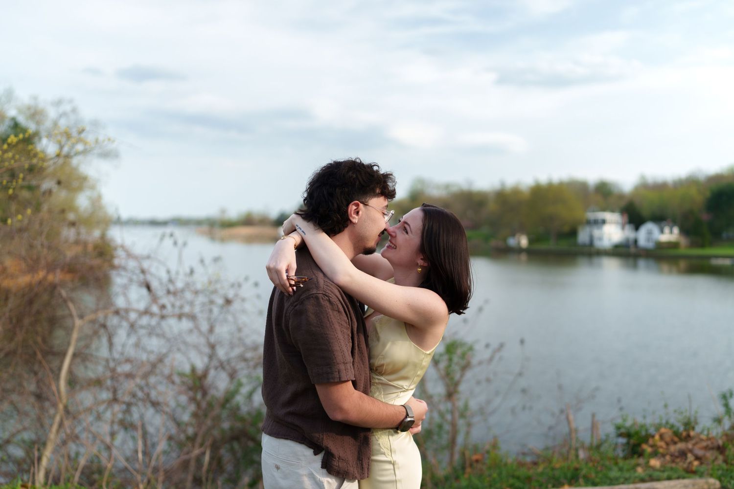 Happy couple embracing and kissing by a scenic lake with trees in background.