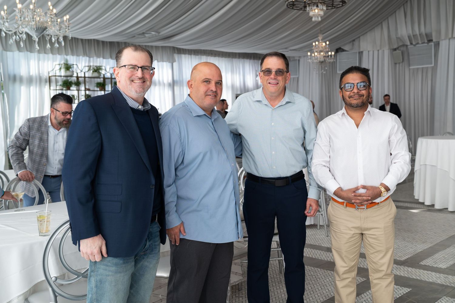 Four men pose together smiling at an elegant white decorated event venue.