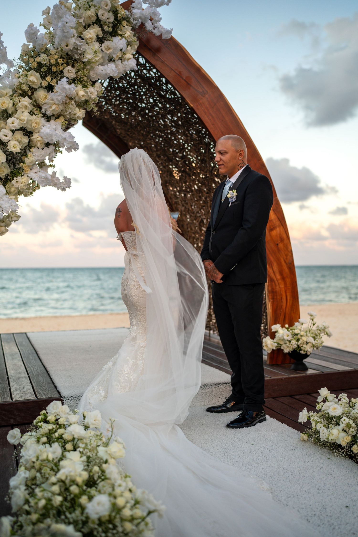 Bride and groom exchange vows under wooden arch adorned with white flowers on tropical beach at sunset.