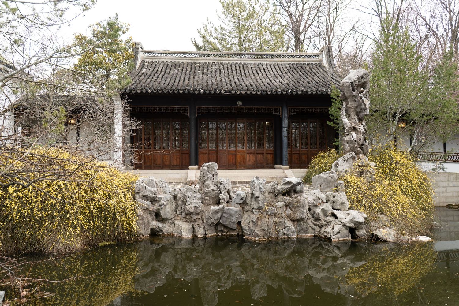 Traditional Chinese garden pavilion with wooden doors, rocky landscape, and calm reflective pond.