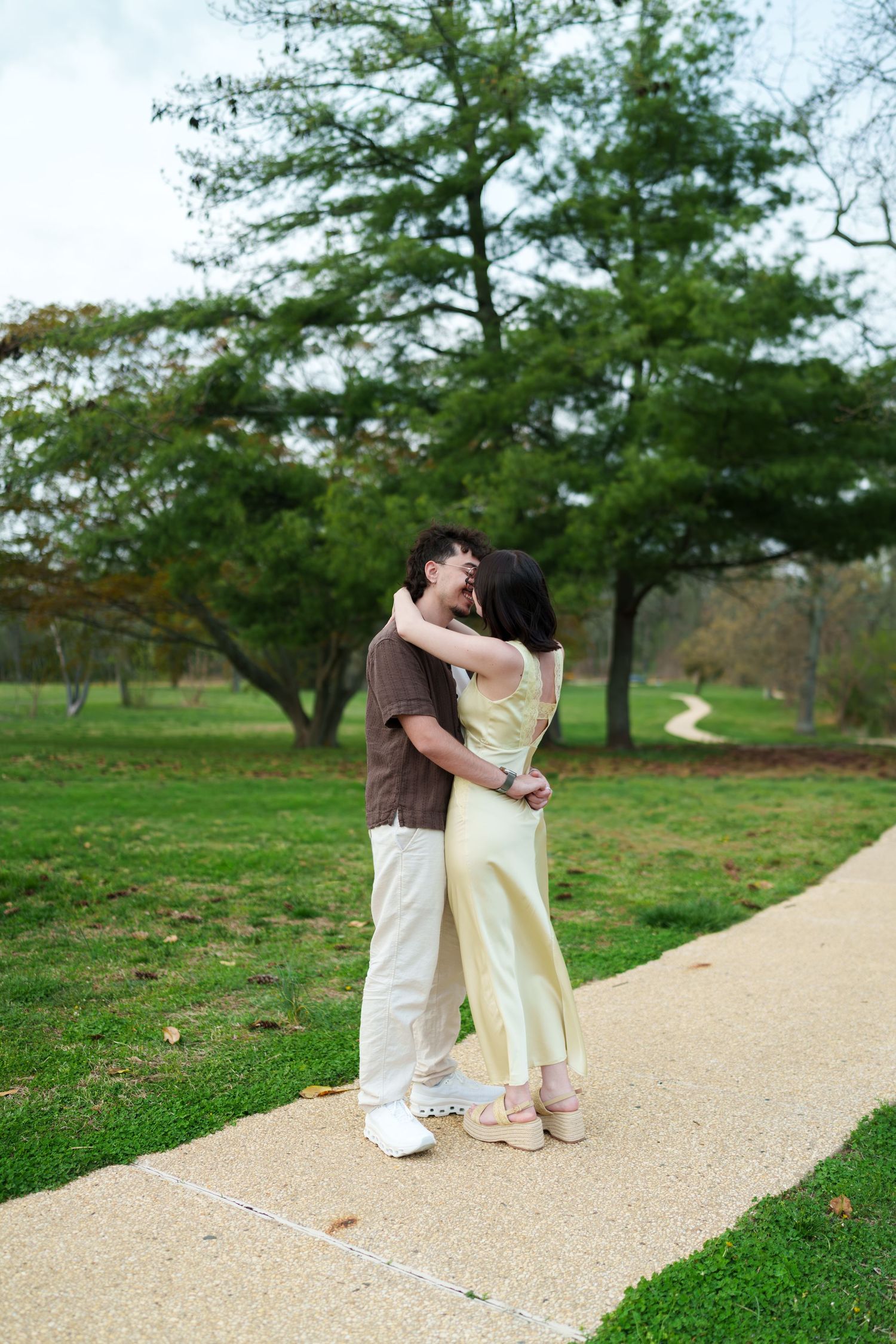 Engaged couple sharing a kiss on a park pathway surrounded by lush green trees.