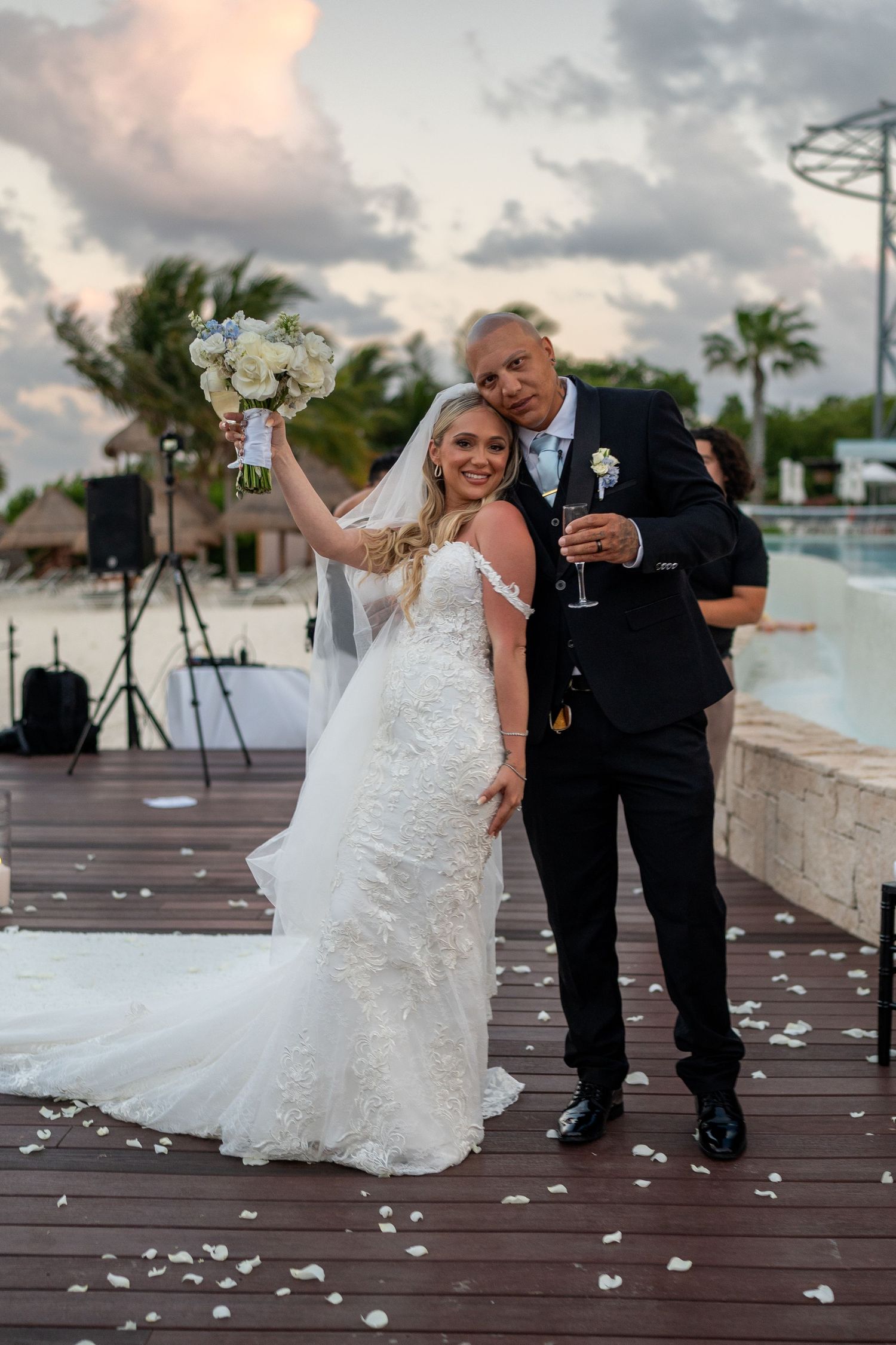 Bride and groom pose joyfully on a waterfront deck, bride raising bouquet, groom holding champagne at sunset wedding.