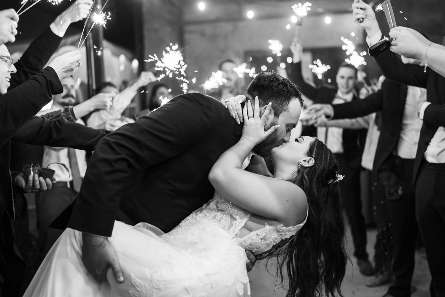 Black and white photo of newlyweds kissing during sparkler exit at wedding reception.