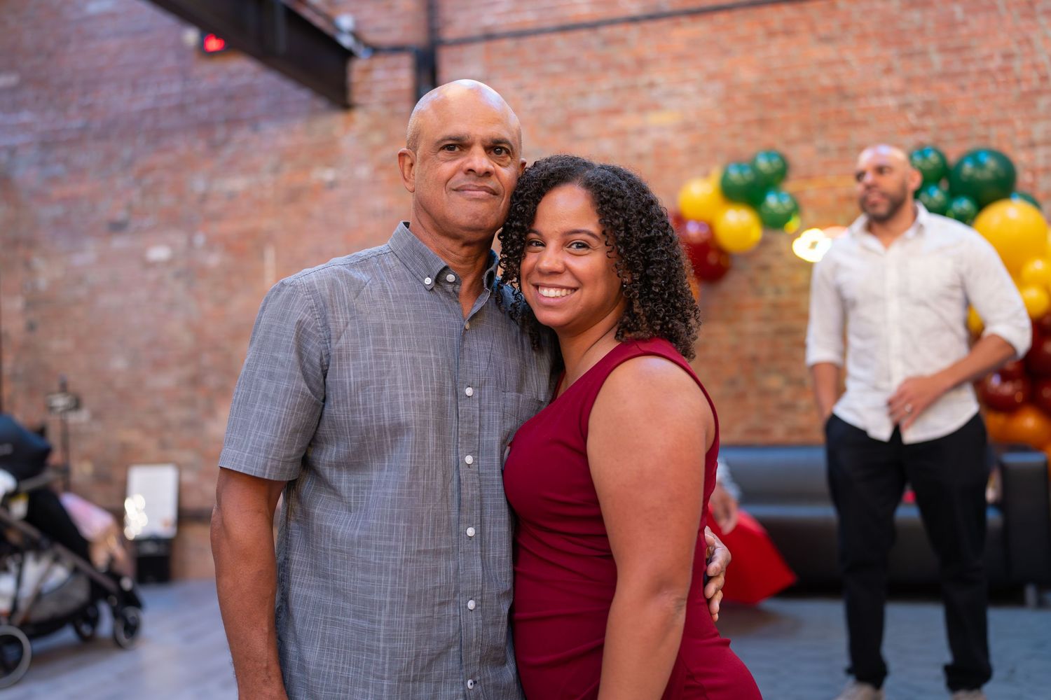 A couple smiles together at an indoor party with colorful balloons and brick wall backdrop.