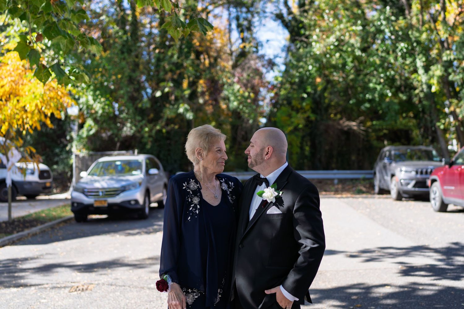 A groom in a black tuxedo shares a tender moment with an older woman outdoors at a wedding.