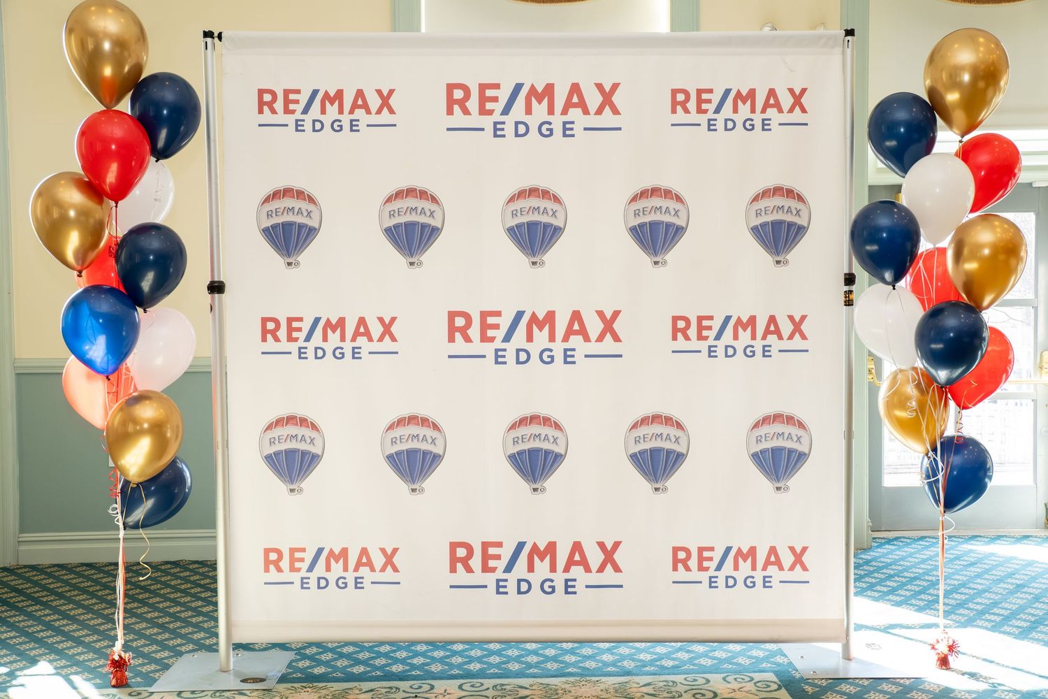 RE/MAX Edge branded step-and-repeat backdrop surrounded by red, blue, and gold balloons at event.
