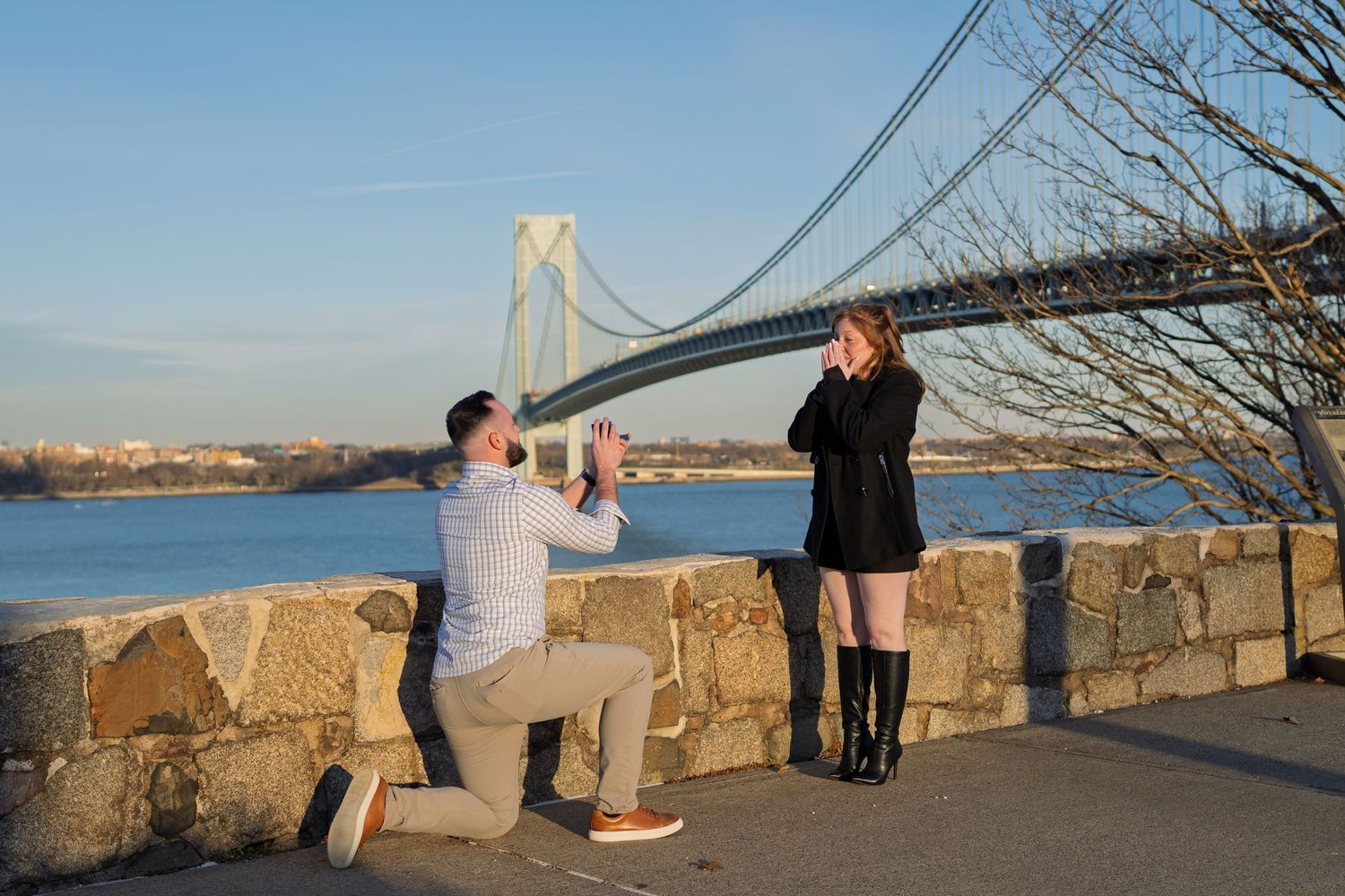 Man kneeling in marriage proposal to woman near river with large suspension bridge in background.