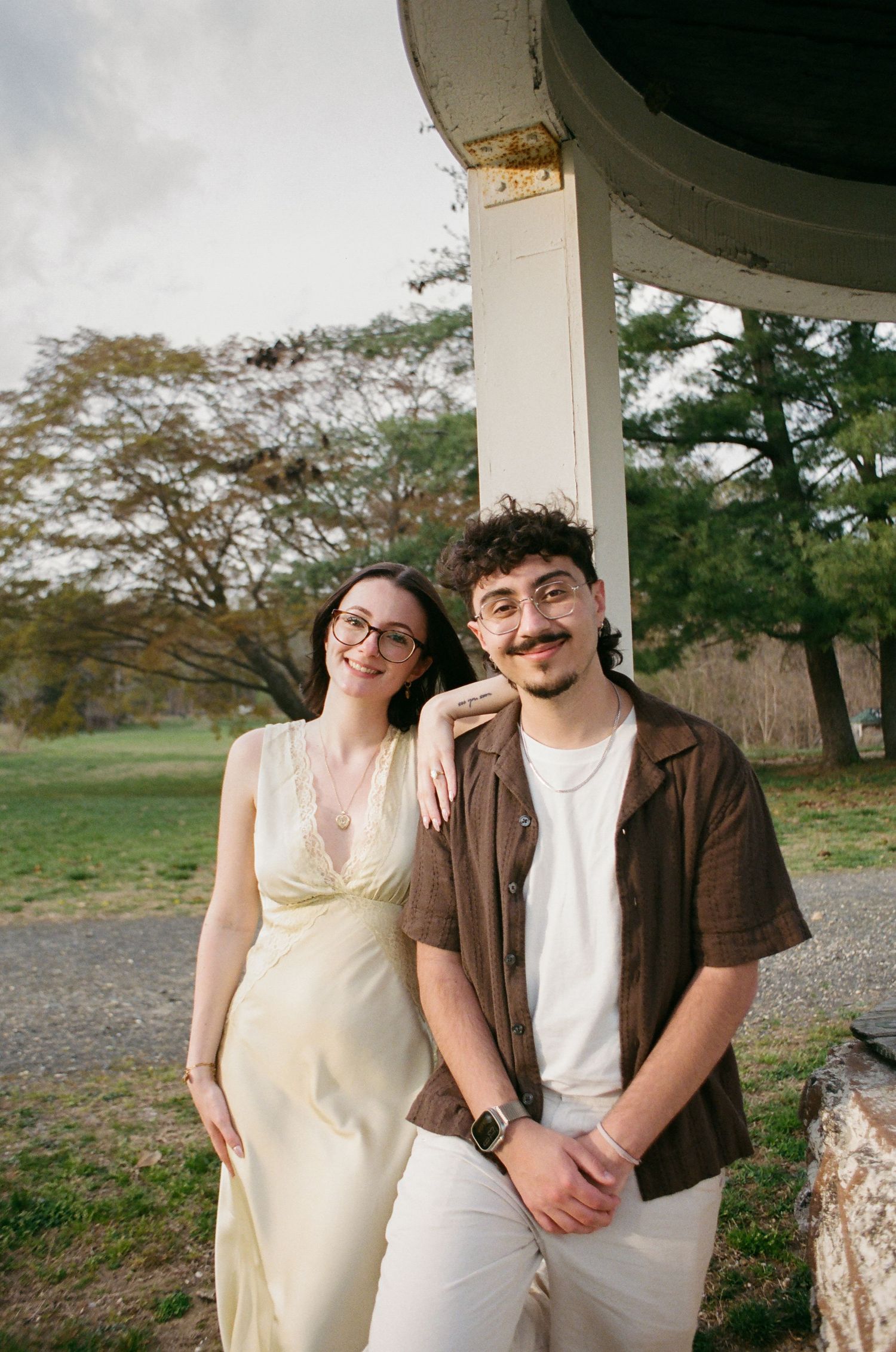 Couple leaning against a white column outdoors, smiling warmly with green trees in the background.