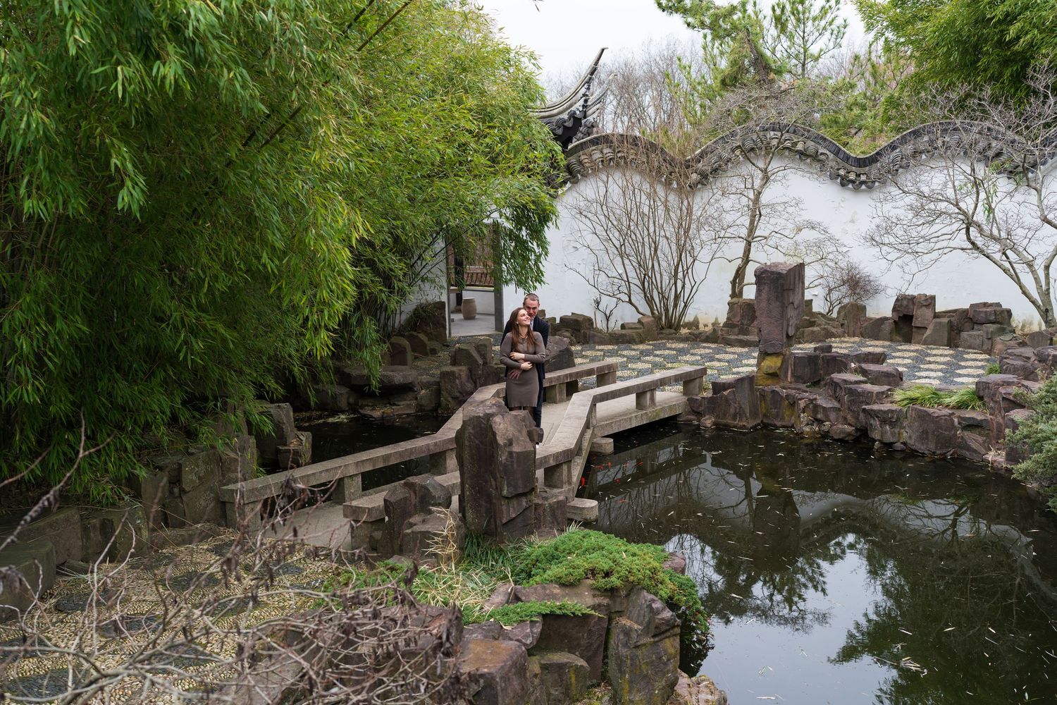 Woman standing on stone path beside tranquil pond in classical Chinese garden with bamboo and white wall.