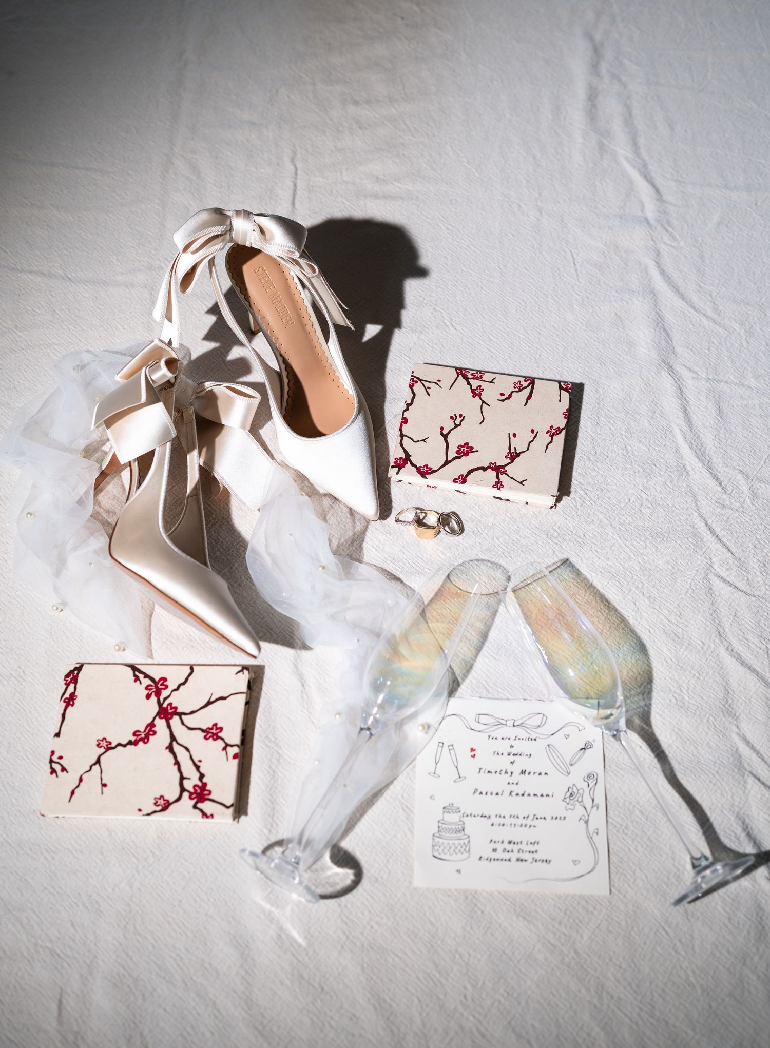 Bridal flat lay with white satin heels, cherry blossom clutch, champagne flutes, veil, rings, and wedding invitation.