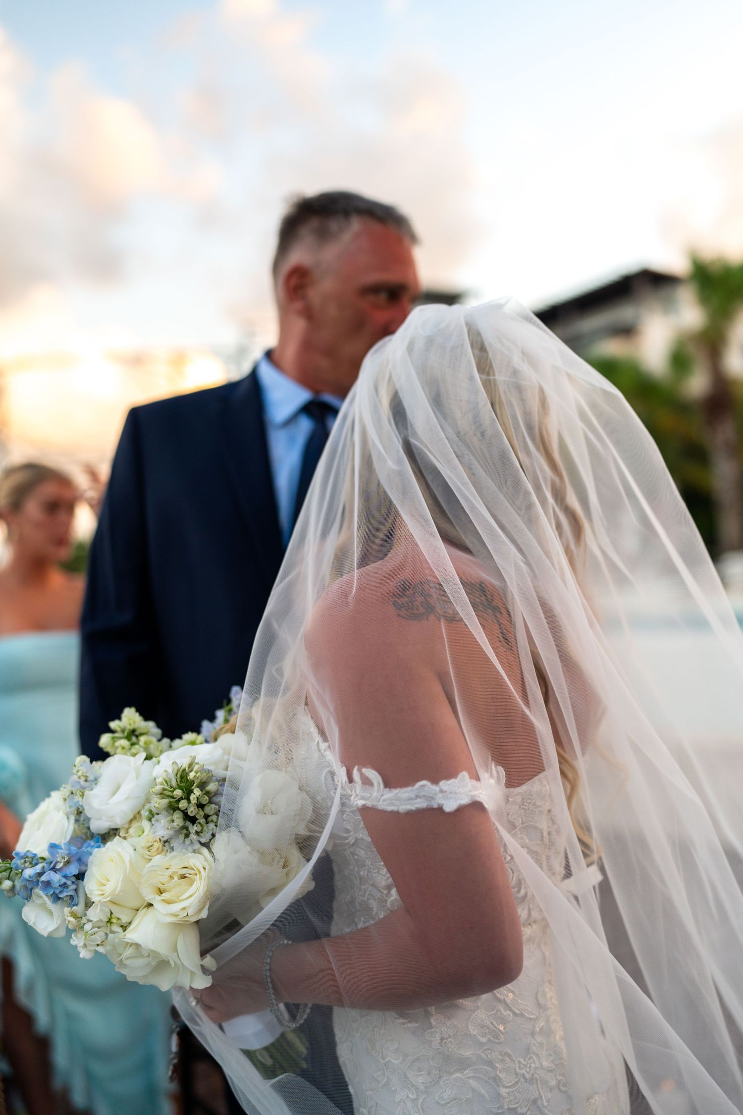 Bride with floral tattoo holding white bouquet under veil during outdoor beach wedding ceremony at sunset.