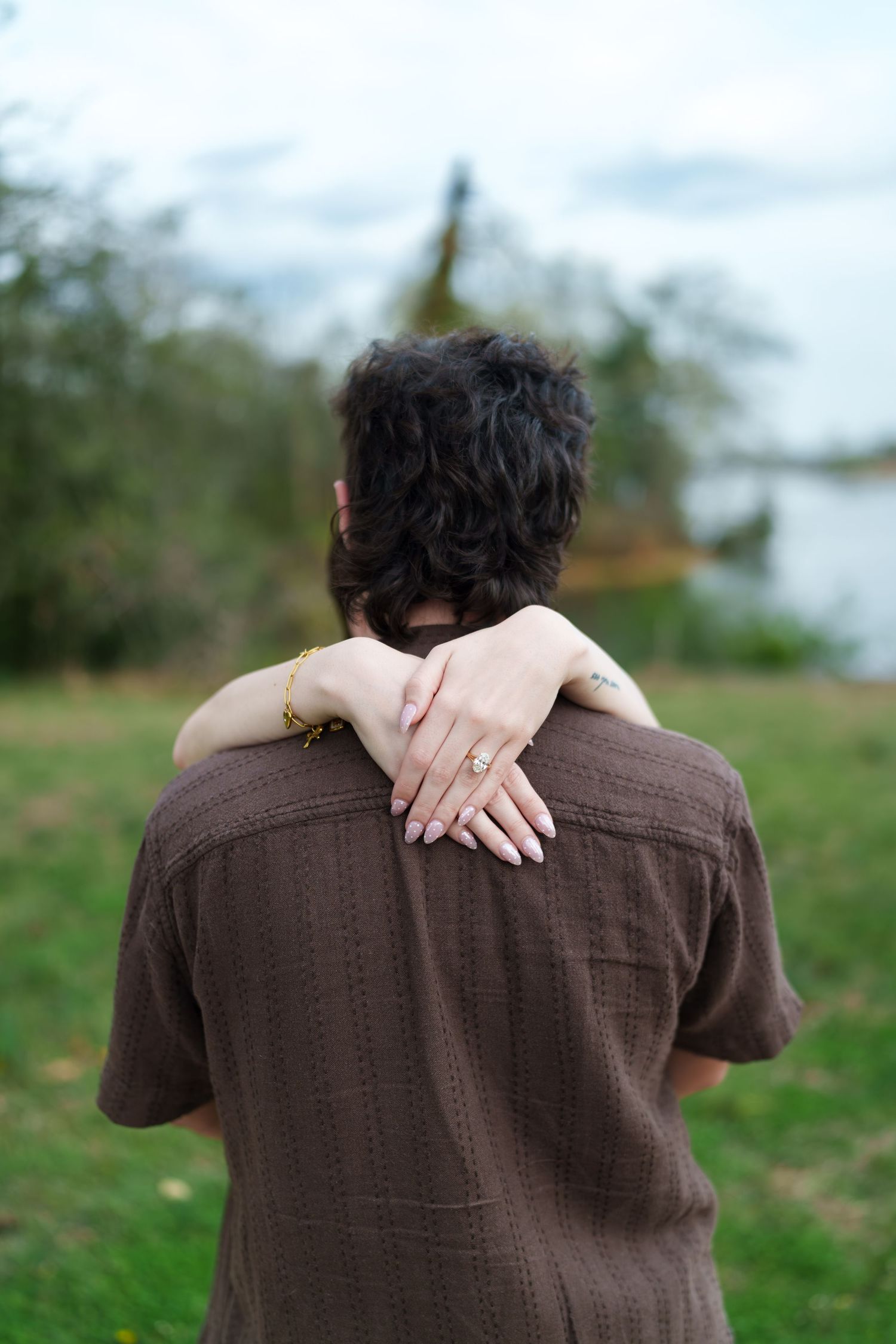 Close-up of woman's hand with engagement ring resting on man's brown shirt back outdoors.
