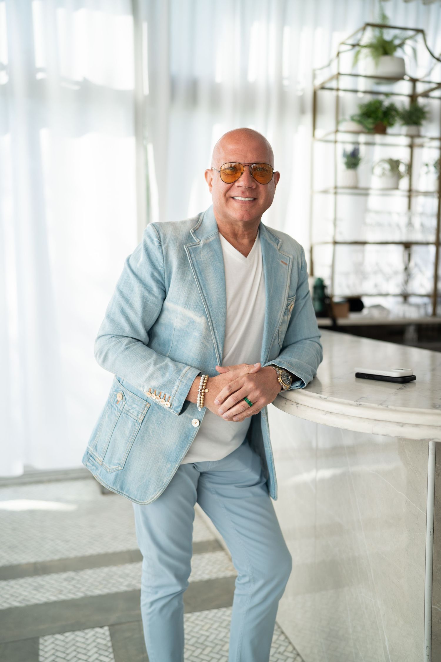 Confident bald man in light blue suit and sunglasses leaning against a marble counter.