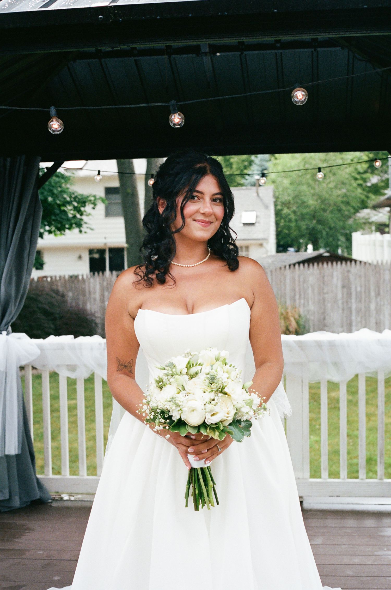 Bride in strapless white gown holding white rose bouquet smiling under gazebo at wedding.