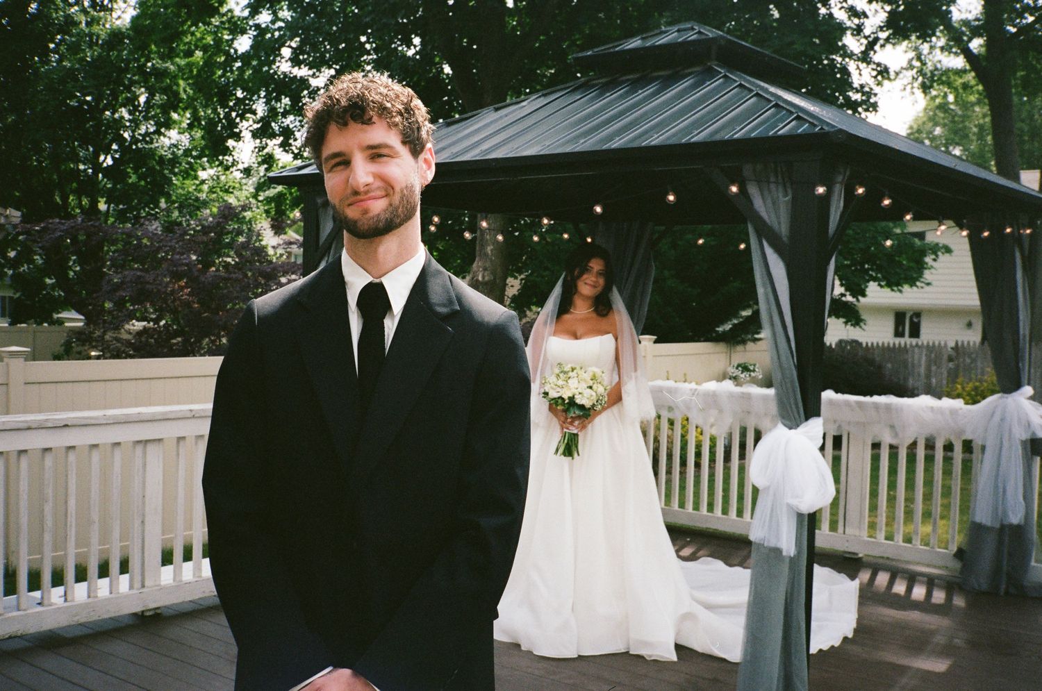 Groom in black suit stands on deck as bride in white gown approaches from behind.