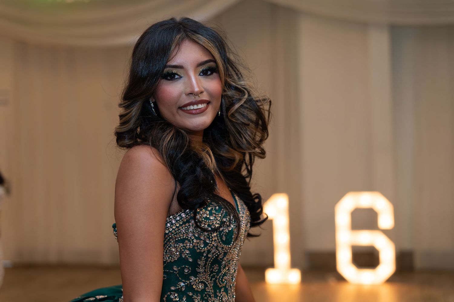 Young woman in green gown smiles at her quinceañera celebration with illuminated 16 sign.