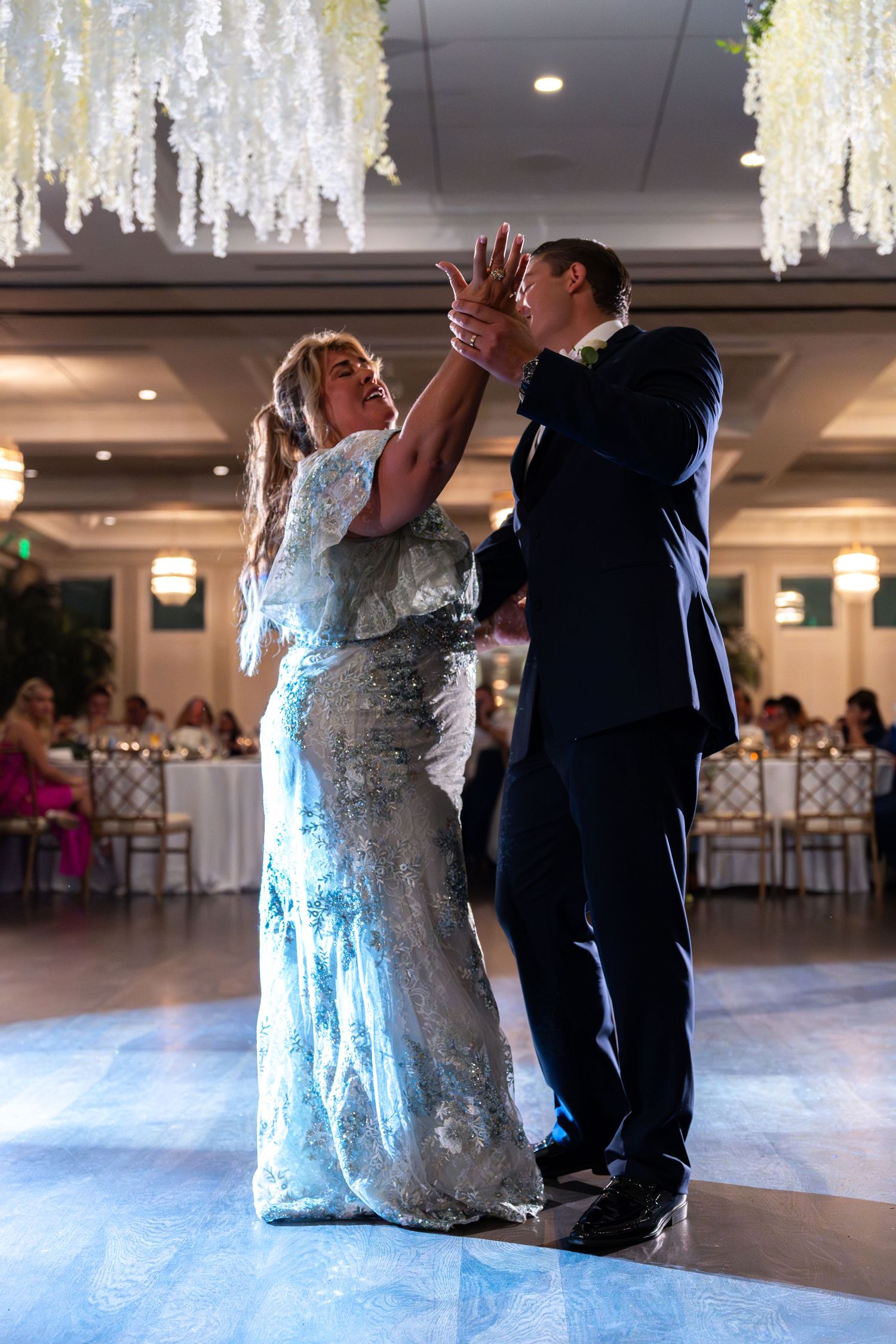Mother and groom share an emotional dance under floral chandelier at wedding reception.