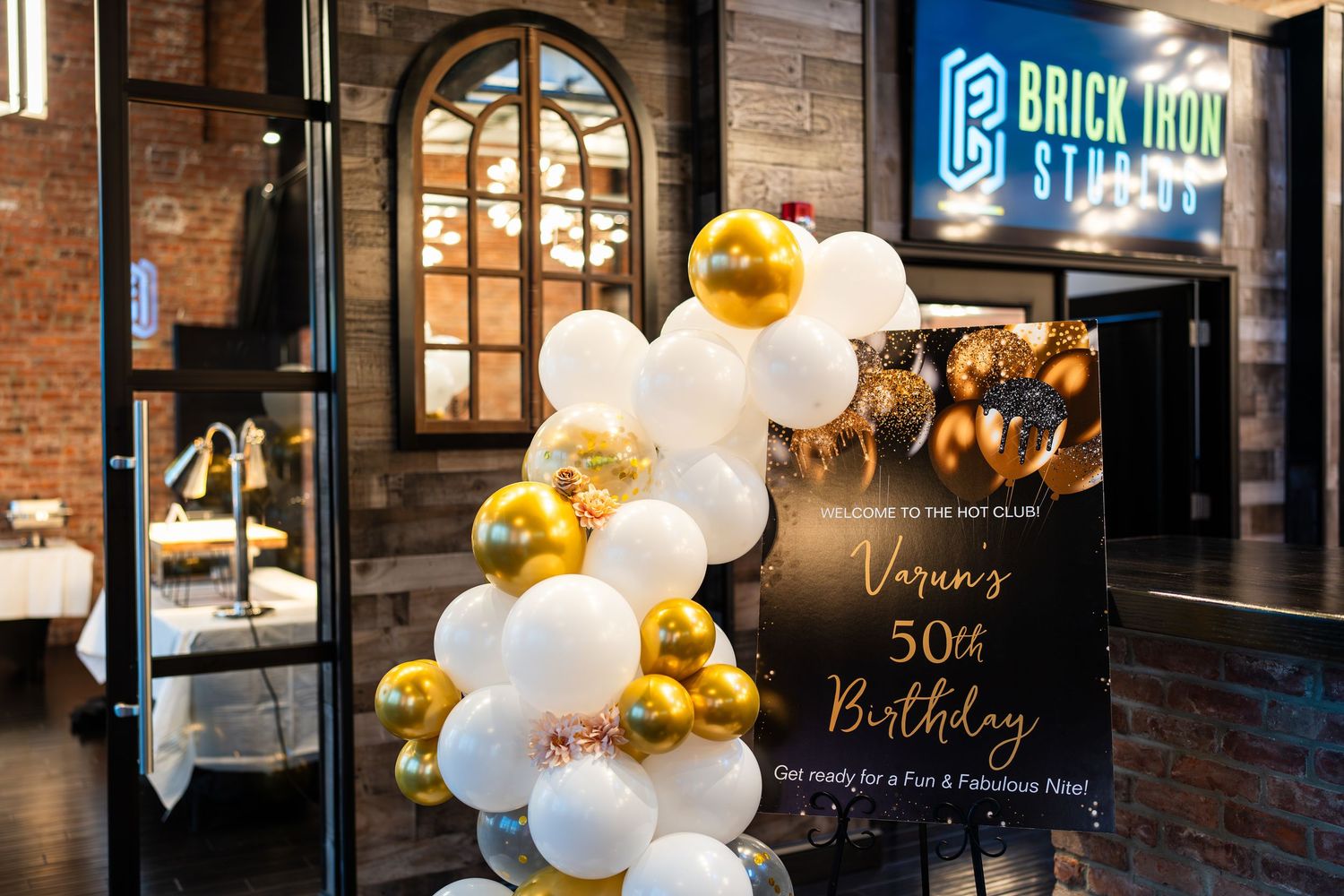 White and gold balloon garland arch beside a 50th birthday welcome sign at Brick Iron Studios.