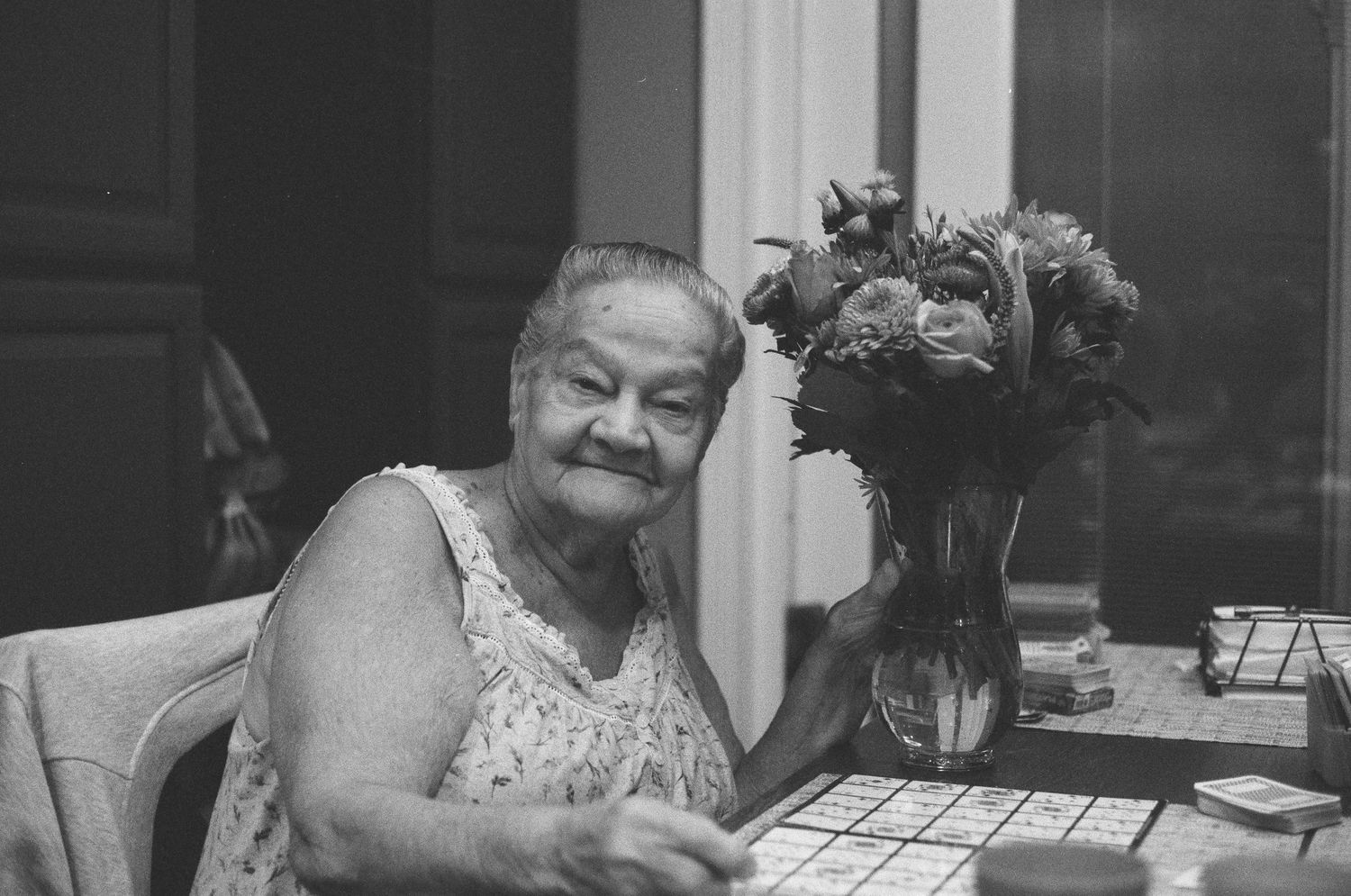 An elderly woman smiles while sitting at a table next to a glass vase of flowers in black and white.