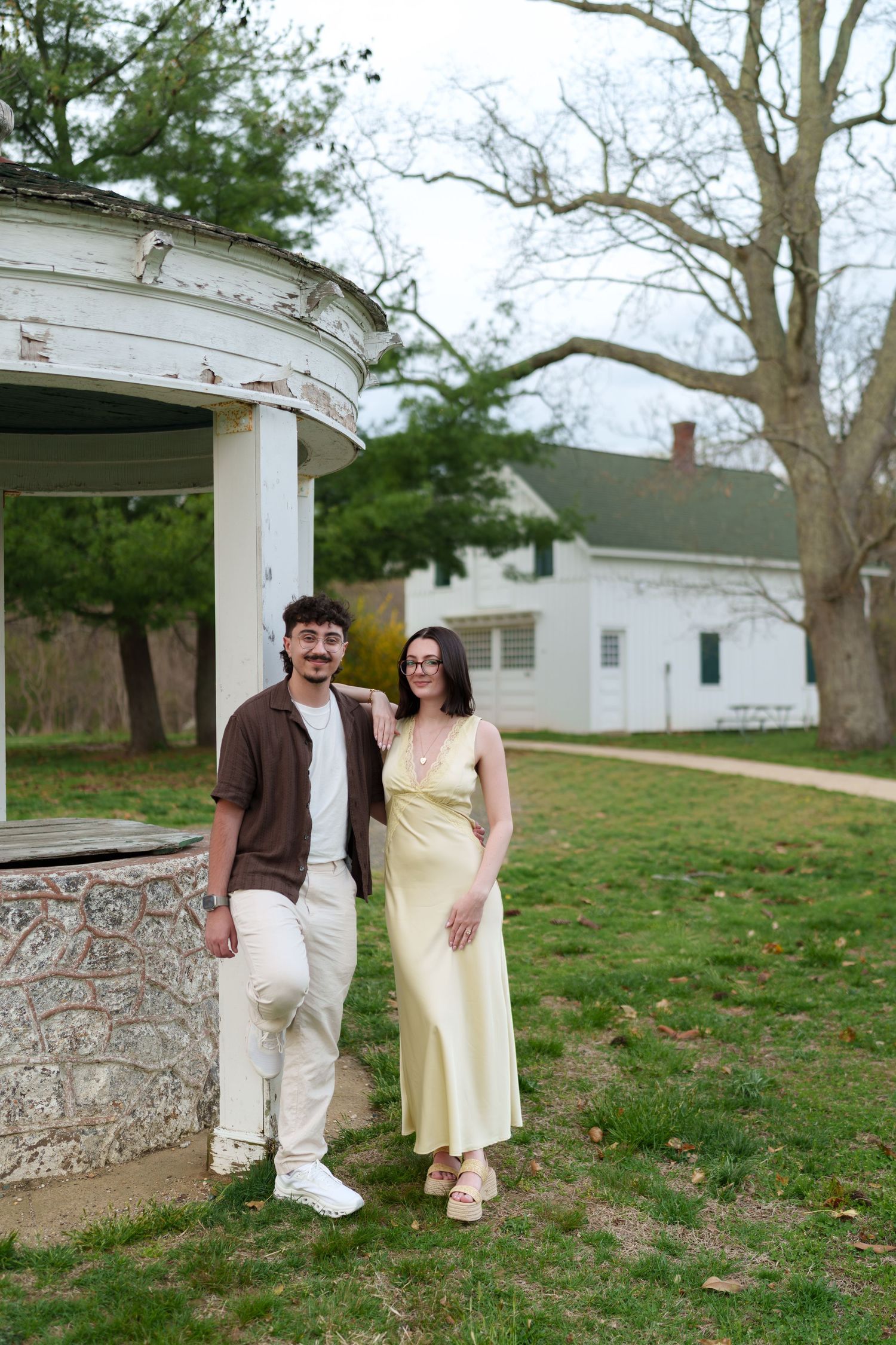 Couple standing together near a stone well and white gazebo in a green park with historic house.