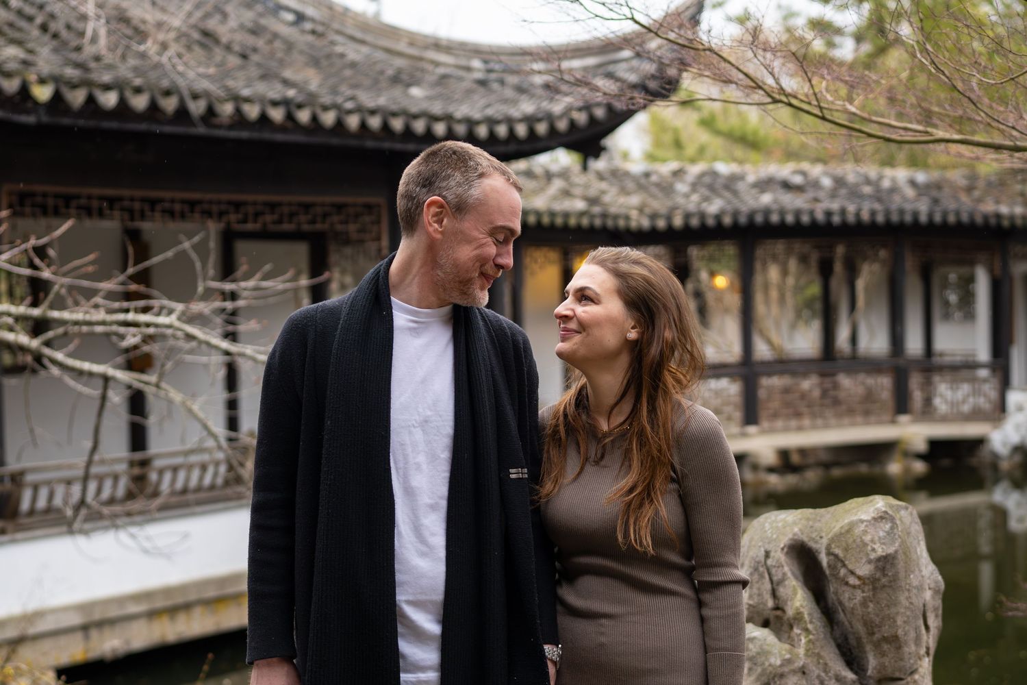 Couple sharing a warm smile beside a peaceful Chinese garden pond with a pagoda backdrop.