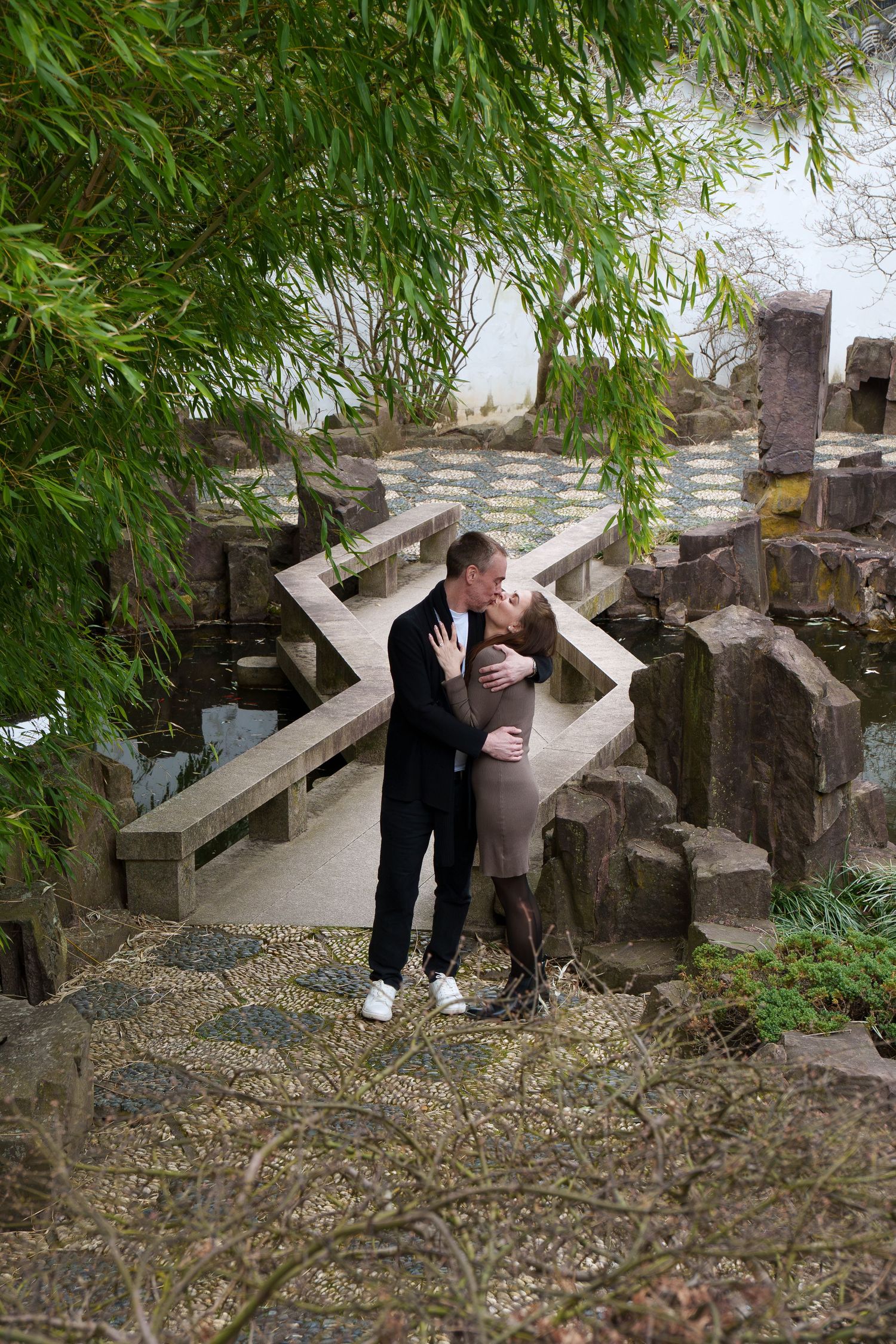 Couple sharing a romantic kiss on a stone path in a lush Asian garden.