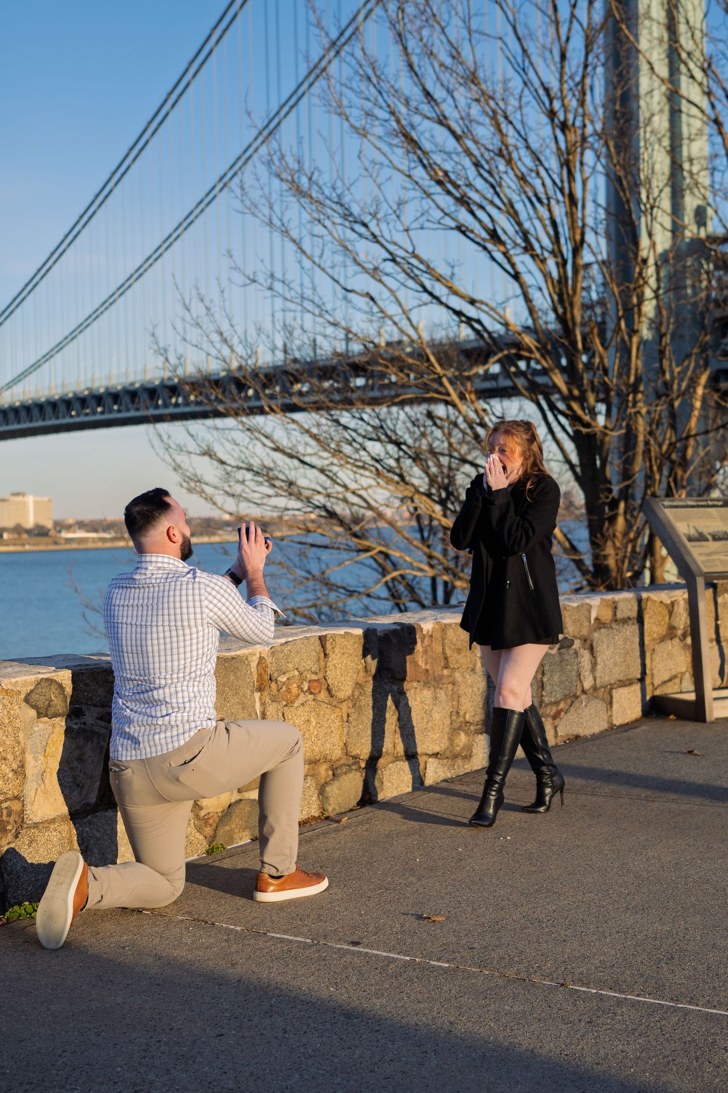 Man proposing on one knee to his girlfriend near a scenic bridge waterfront at sunset.